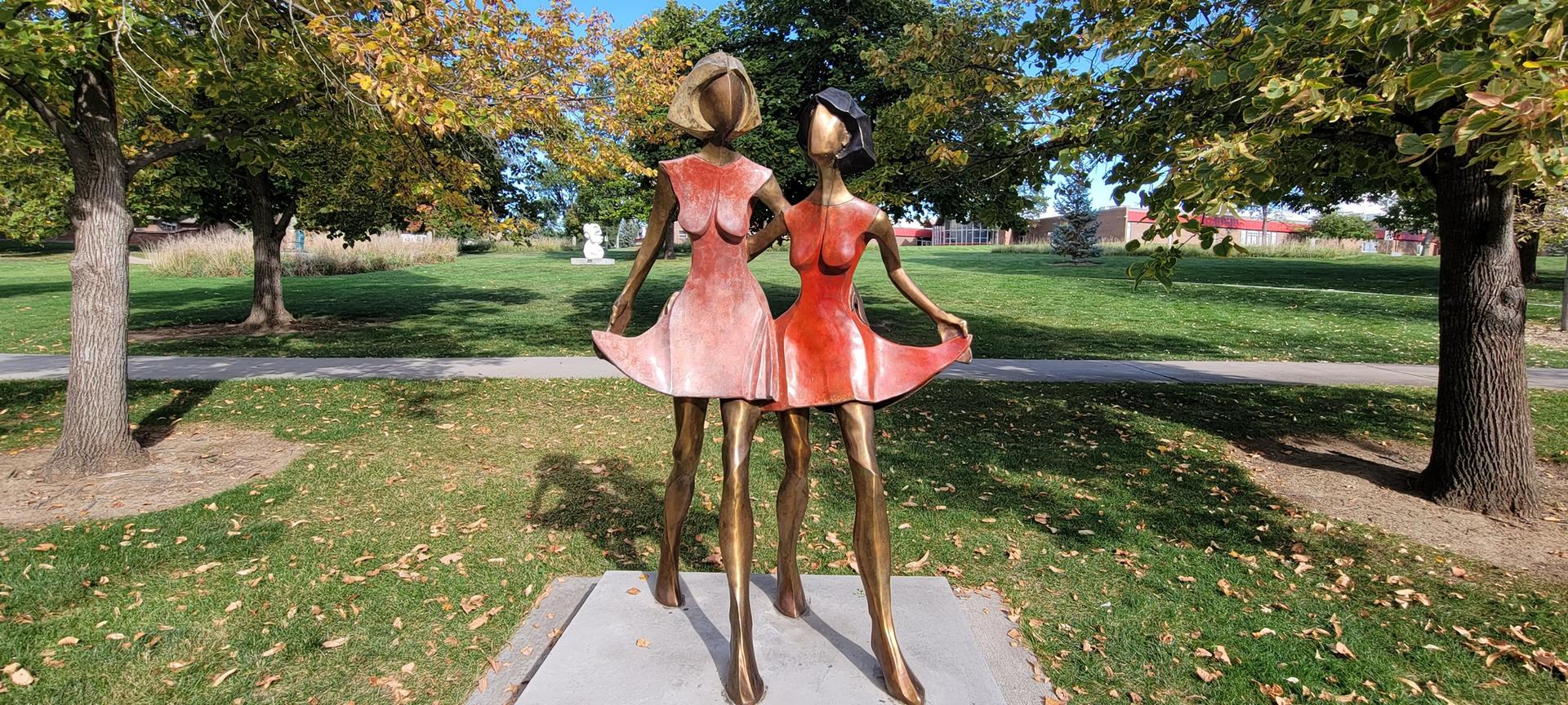 Bronze statue of two women in dresses on a sunny green lawn.