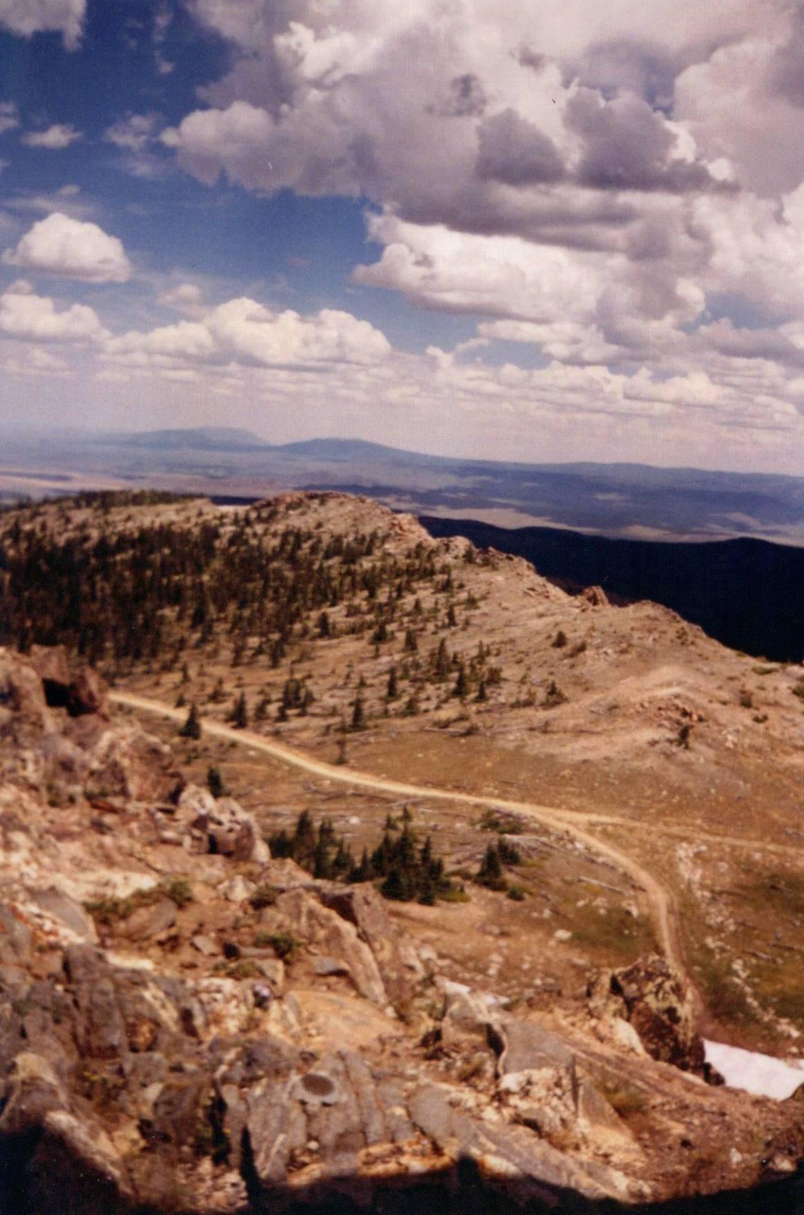 A winding dirt road traverses rocky, treed mountains under a cloudy sky.