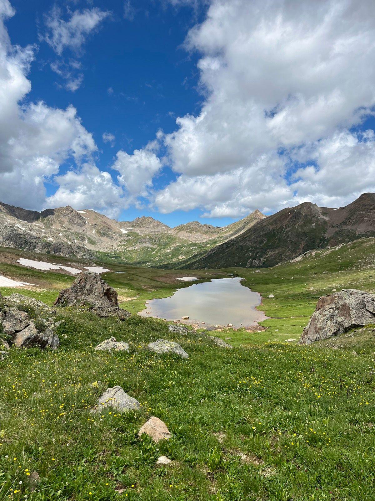 Mountain valley with a small lake, green grass, and rocky peaks under a cloudy sky.