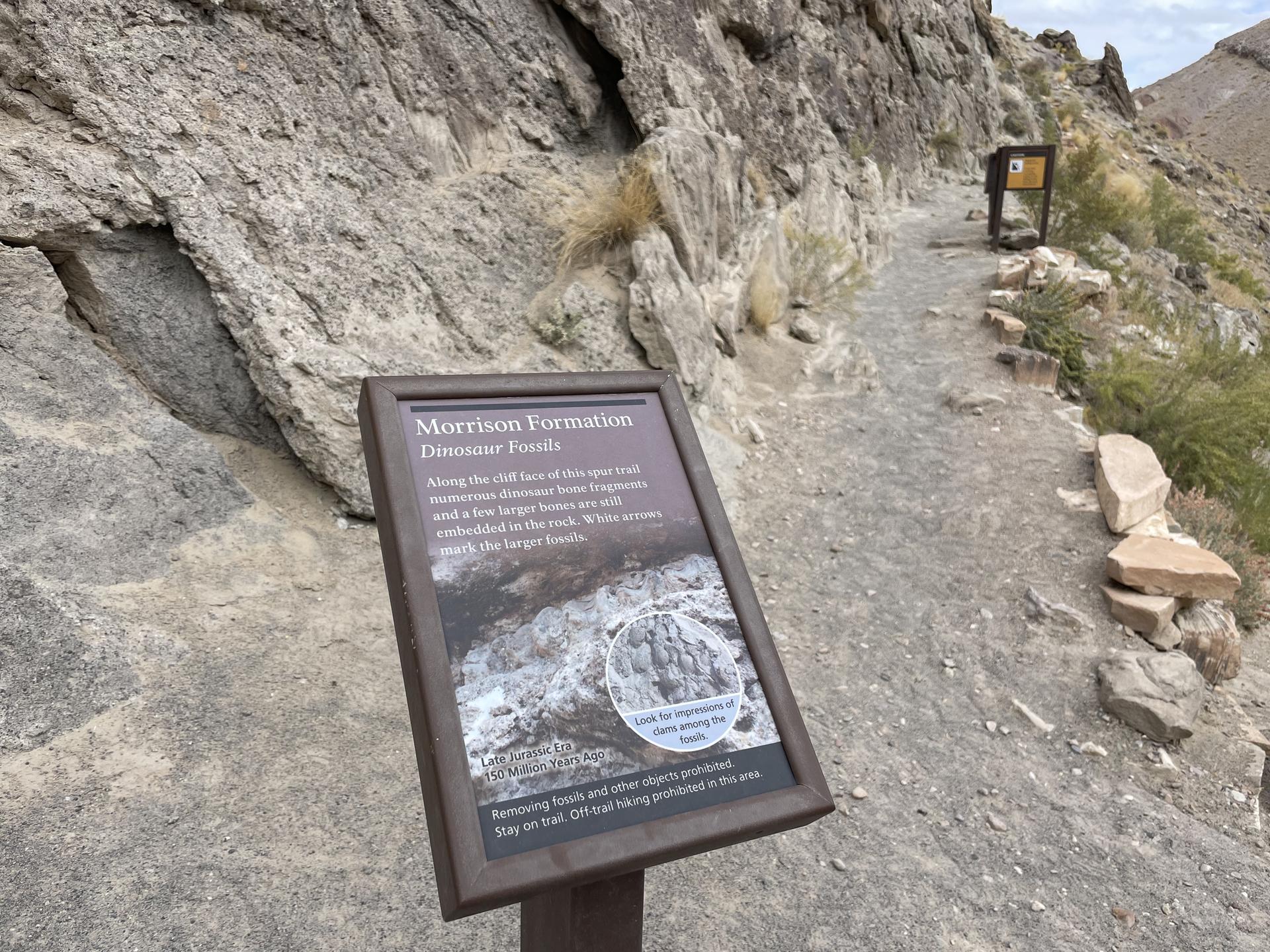 An interpretive sign stands on a dirt path next to a rocky cliff.