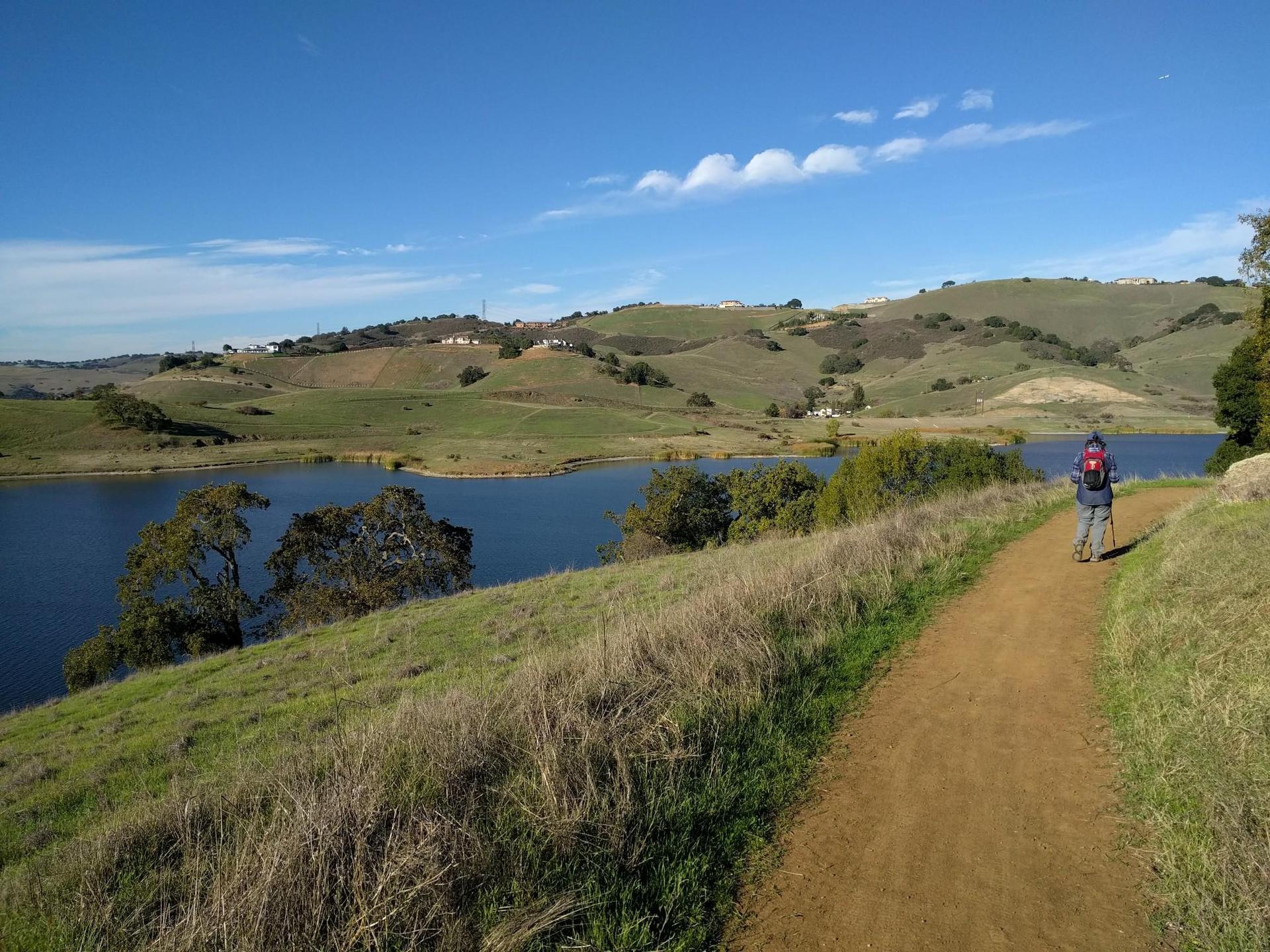 Cyclist on a dirt path beside a lake, surrounded by green hills.