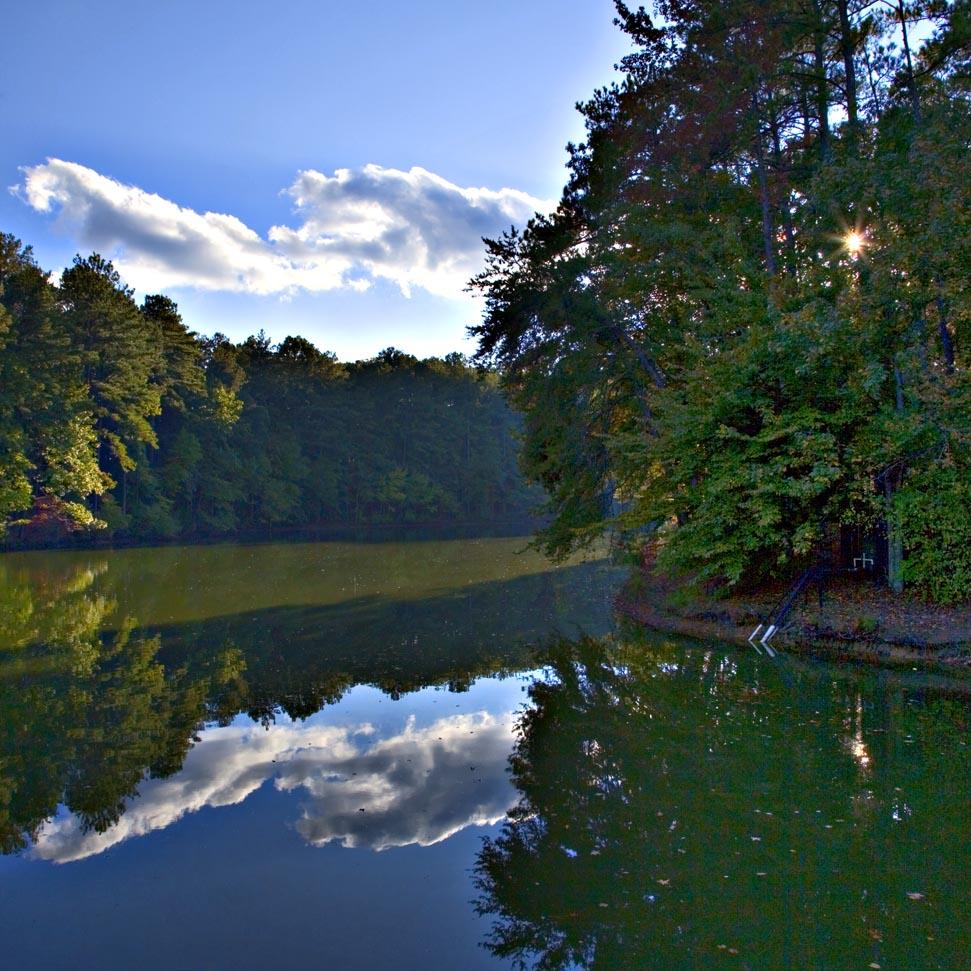 Bright sun peeks through trees over a calm lake with cloud reflections.