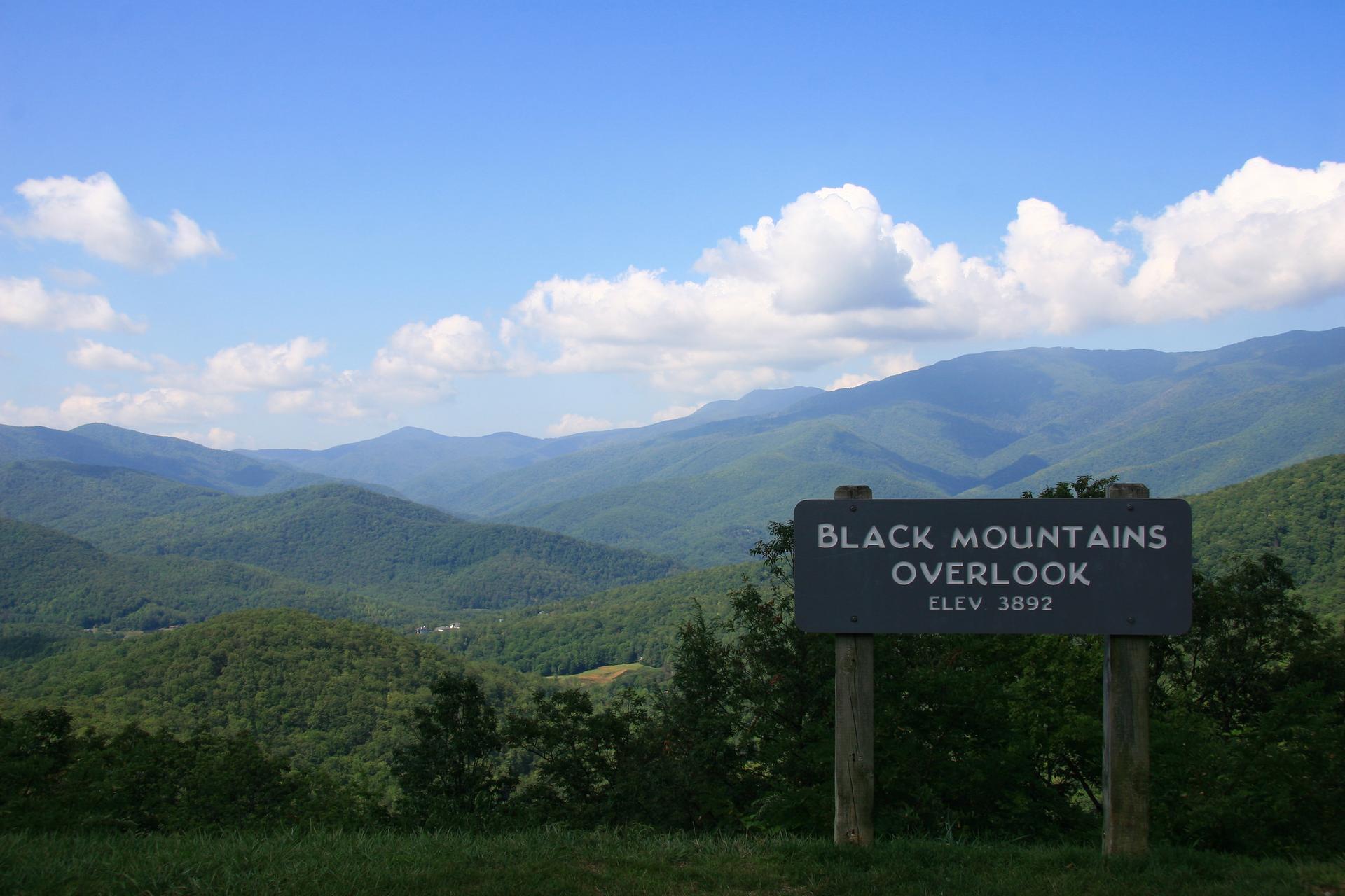 A wooden sign in front of a vast, green mountain range under blue sky.