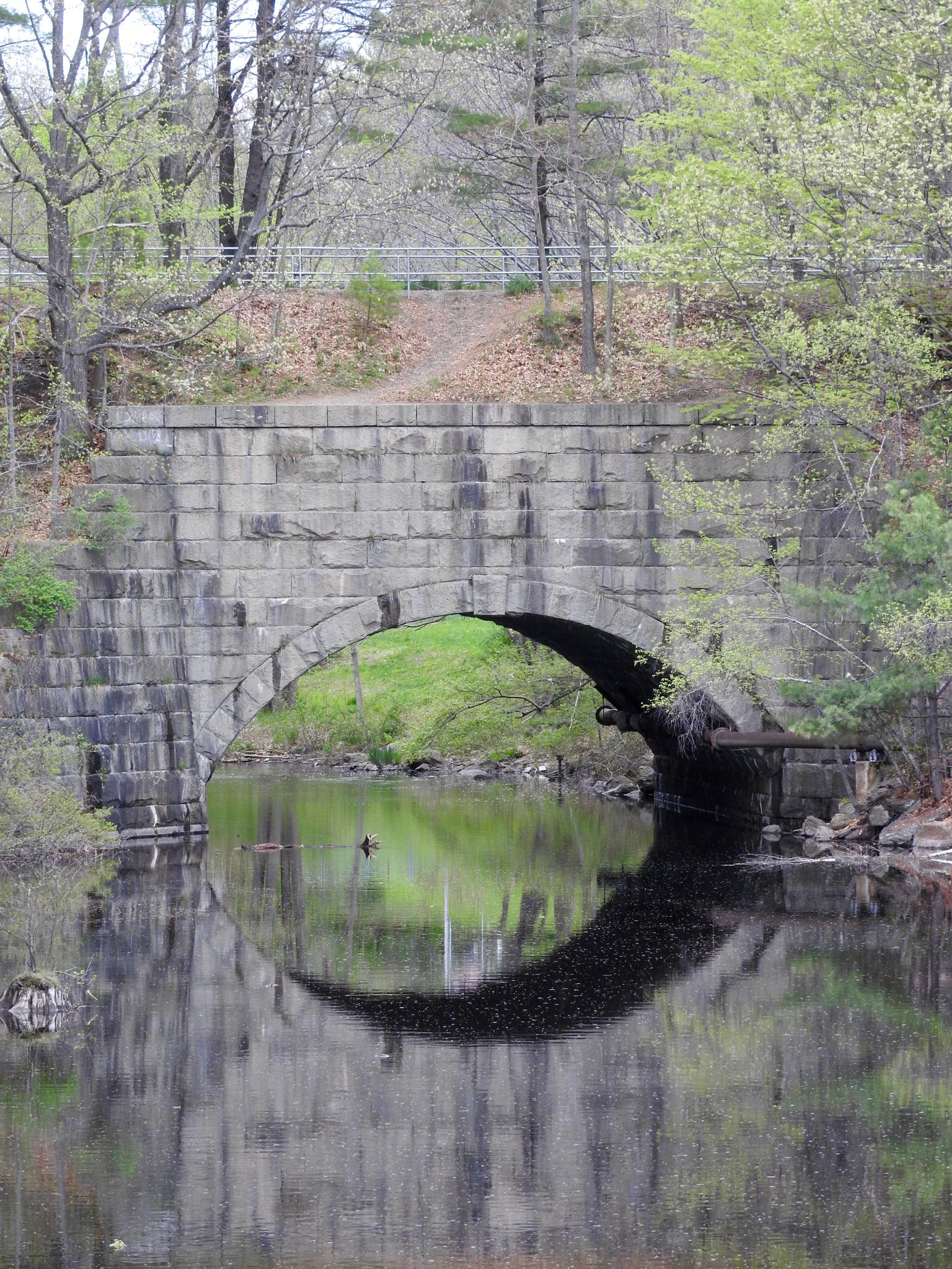 Stone arch bridge over calm water, reflecting trees and sky on an overcast day.