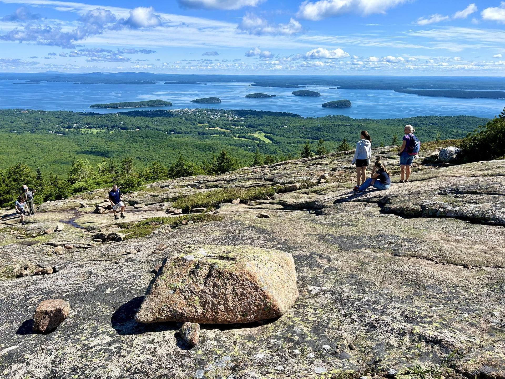 Hikers on a sunny, rocky mountain top overlooking a vast bay with islands.