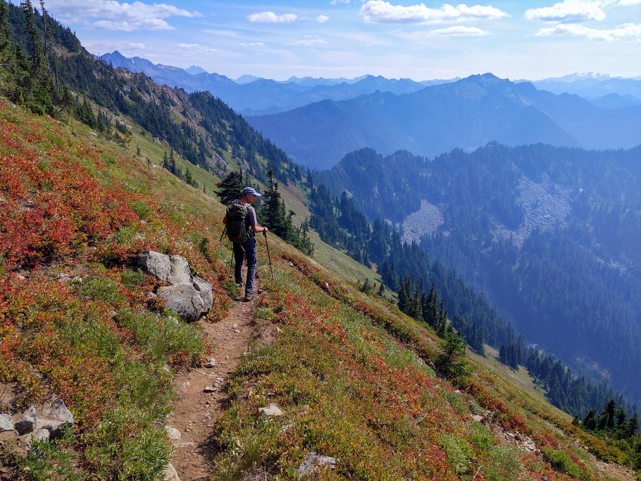 Hiker on a steep mountain trail with colorful foliage and distant peaks.