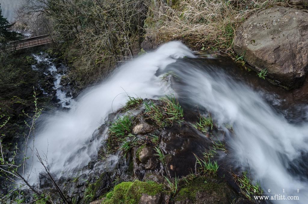 Rushing waterfall cascades over mossy rocks and green foliage, with a bridge below.