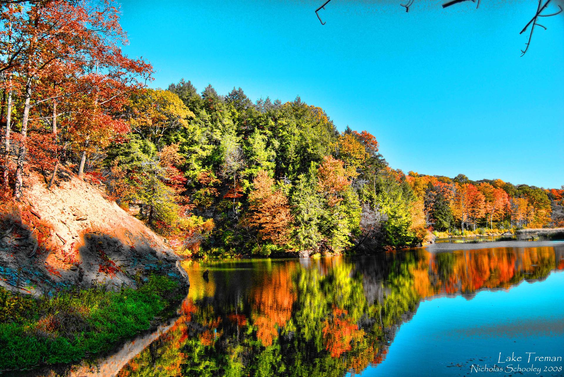 Vibrant autumn trees reflect in a calm lake under a bright blue sky.
