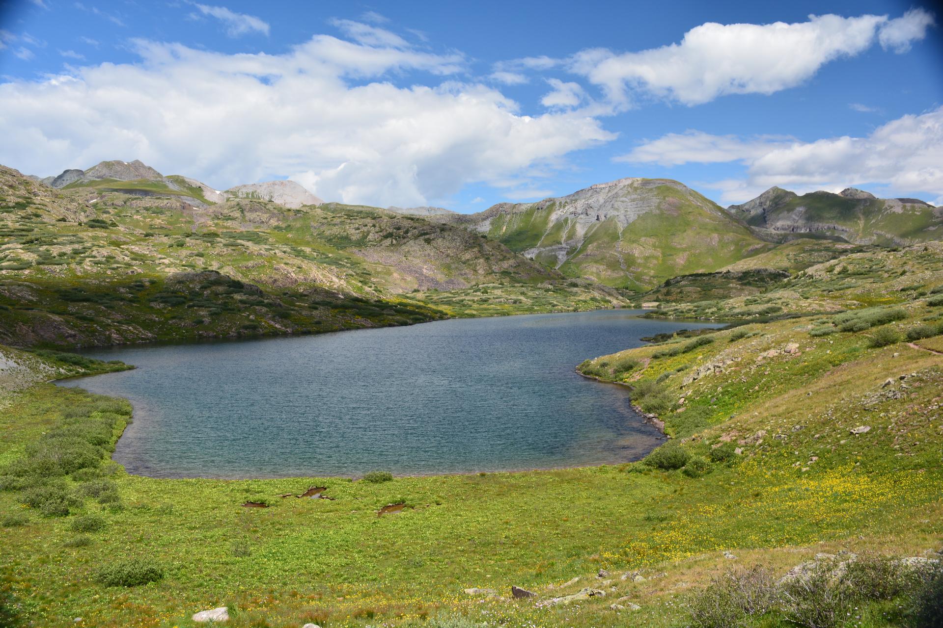 Large mountain lake nestled among green, rolling hills and distant peaks under a cloudy sky.