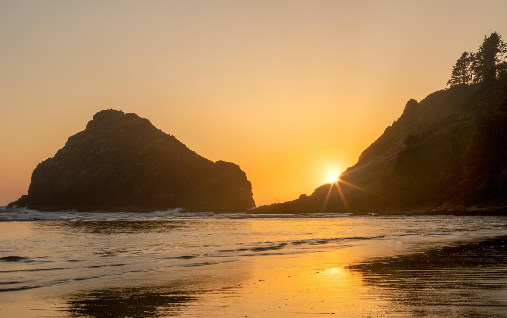 Golden sunset over the ocean, reflecting on the wet sand between two large rock formations.