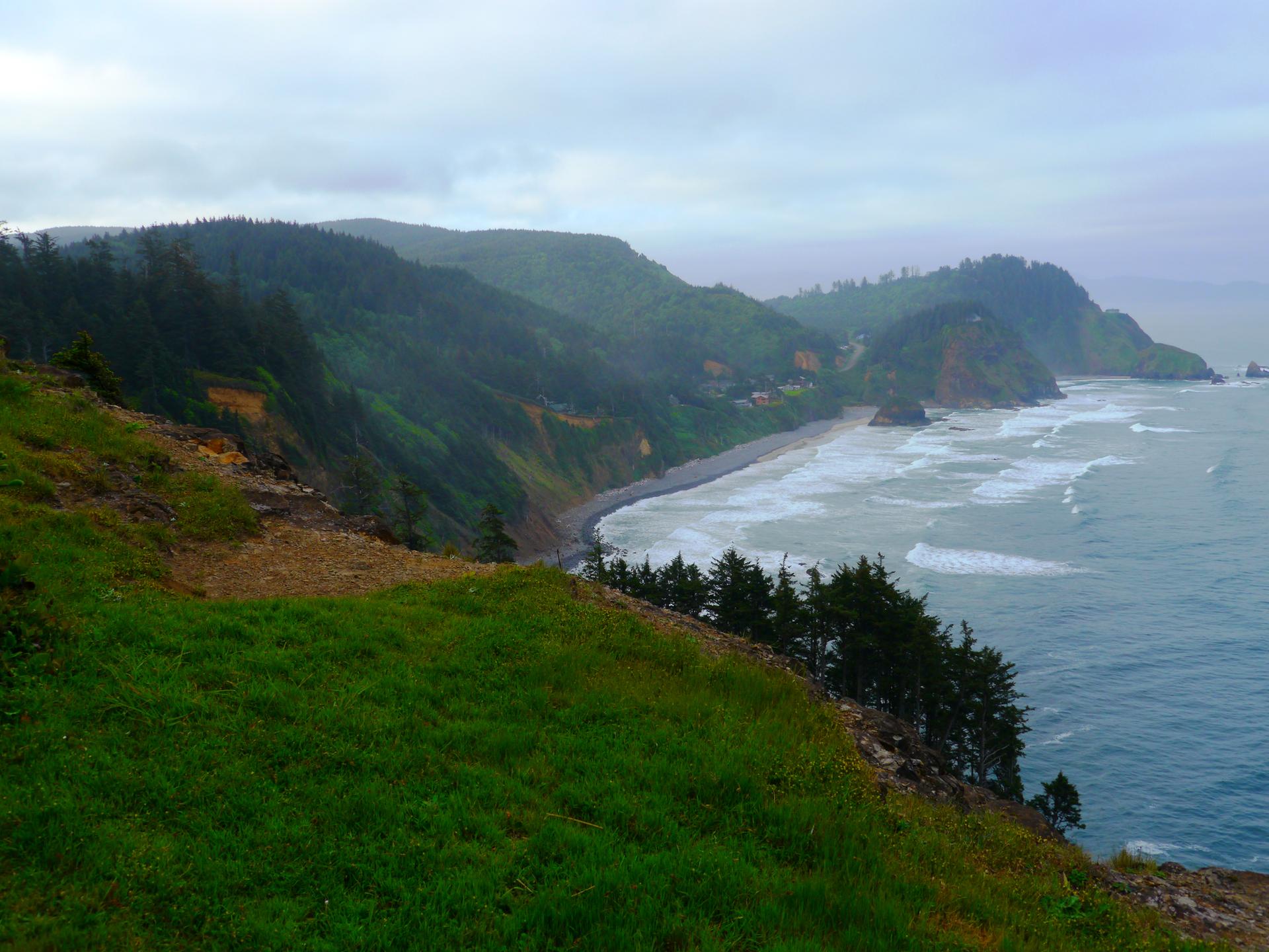 Coastal cliffs covered in green trees overlooking a wavy ocean and sandy beach.