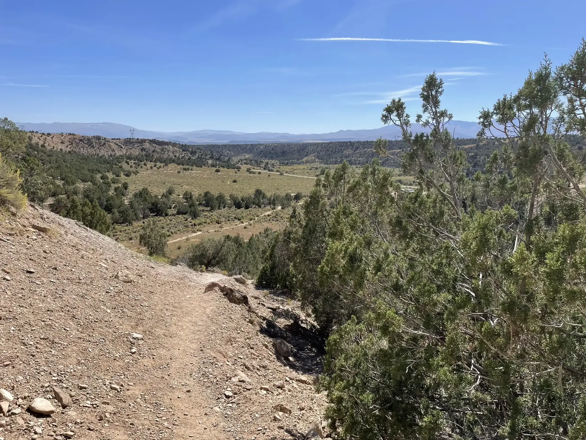 Dirt trail descends into a green valley surrounded by arid hills and juniper trees.