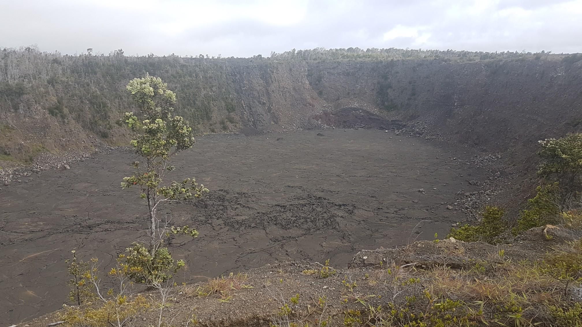 A vast, dark crater with steep walls and sparse trees under an overcast sky. - Halema‘uma‘u and Keanakāko‘i Craters