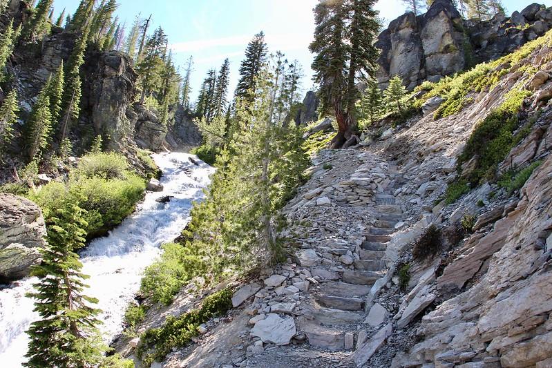 A rocky trail with steps ascends beside a rushing river in a sunlit canyon.