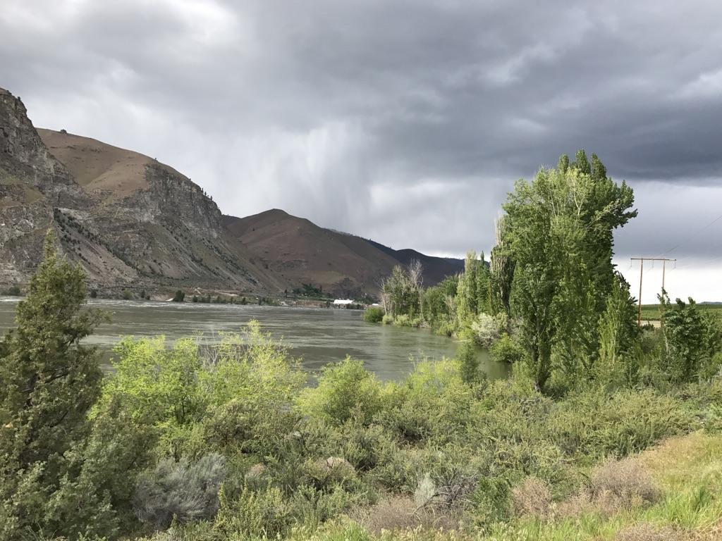 Stormy sky over a wide river, flanked by mountains and green trees.