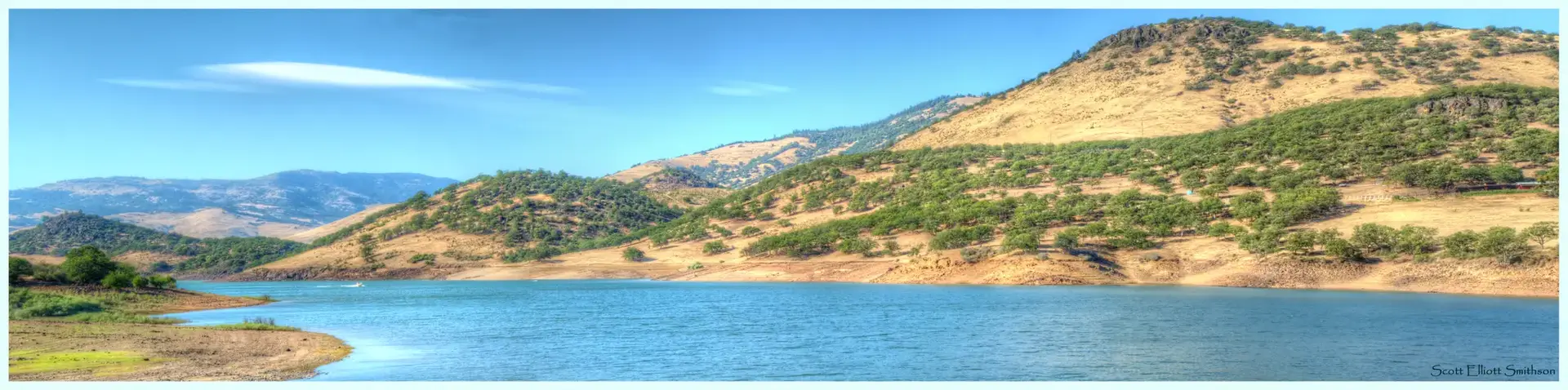 Panoramic view of a blue lake surrounded by dry, tree-covered hills.