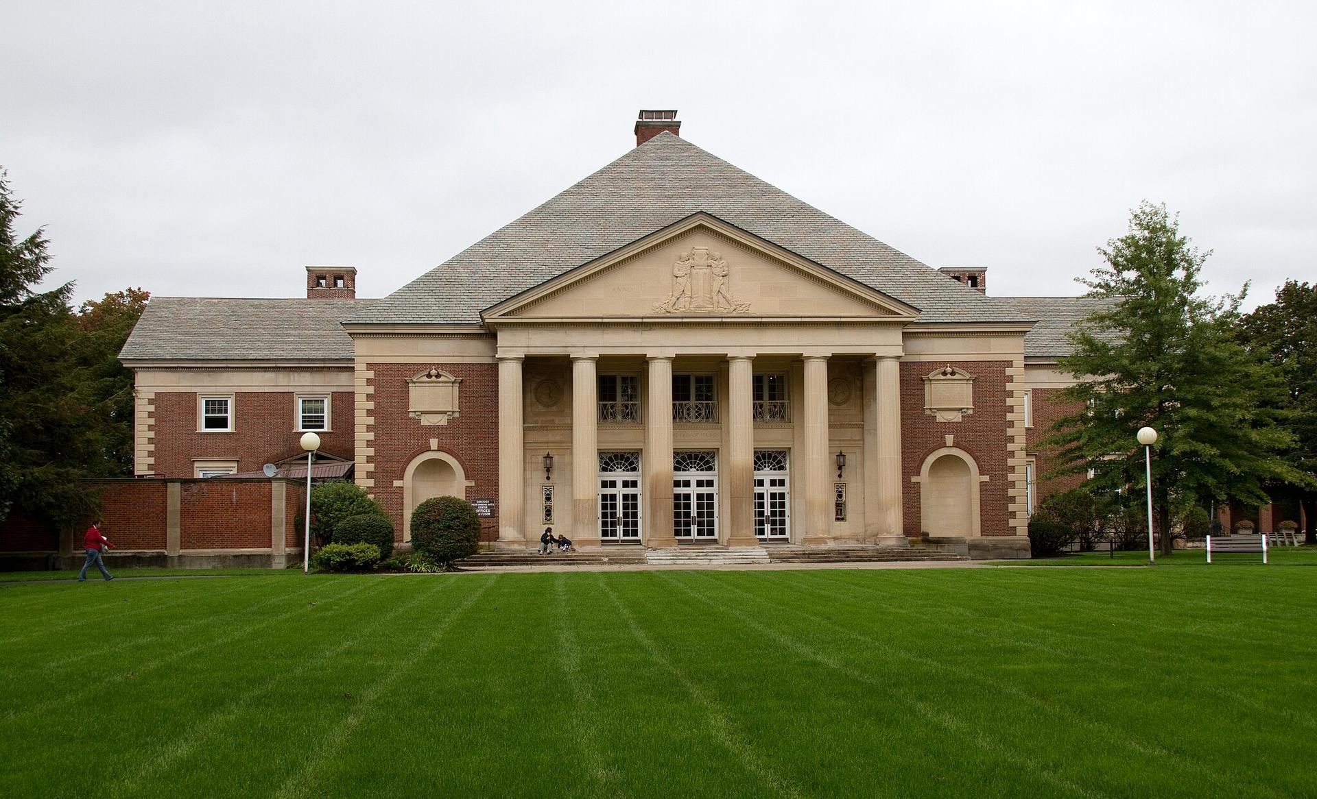 Grand brick building with columns, green lawn, and a person walking under overcast sky.