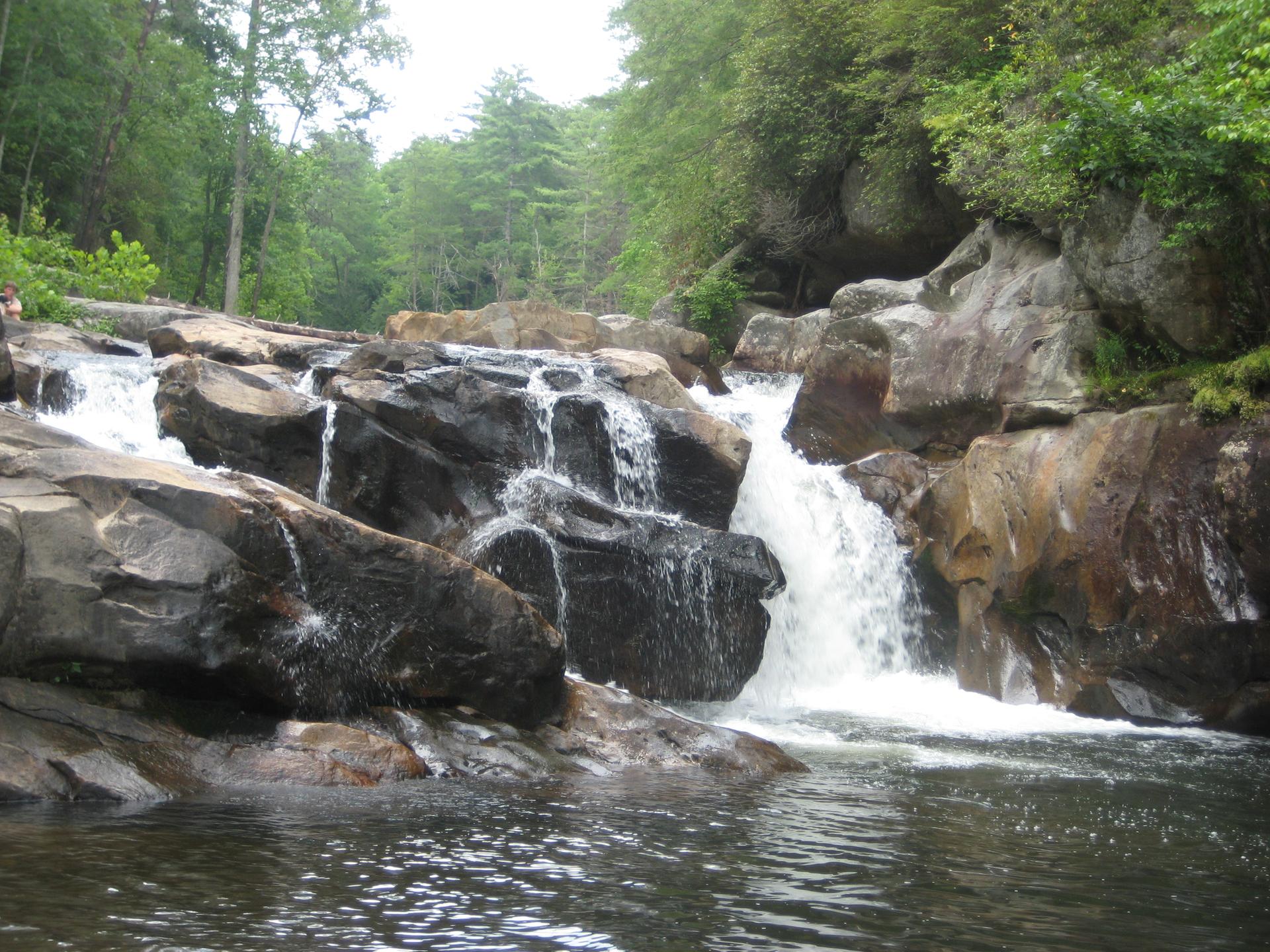 A waterfall cascades over dark rocks into a calm pool, surrounded by green forest.