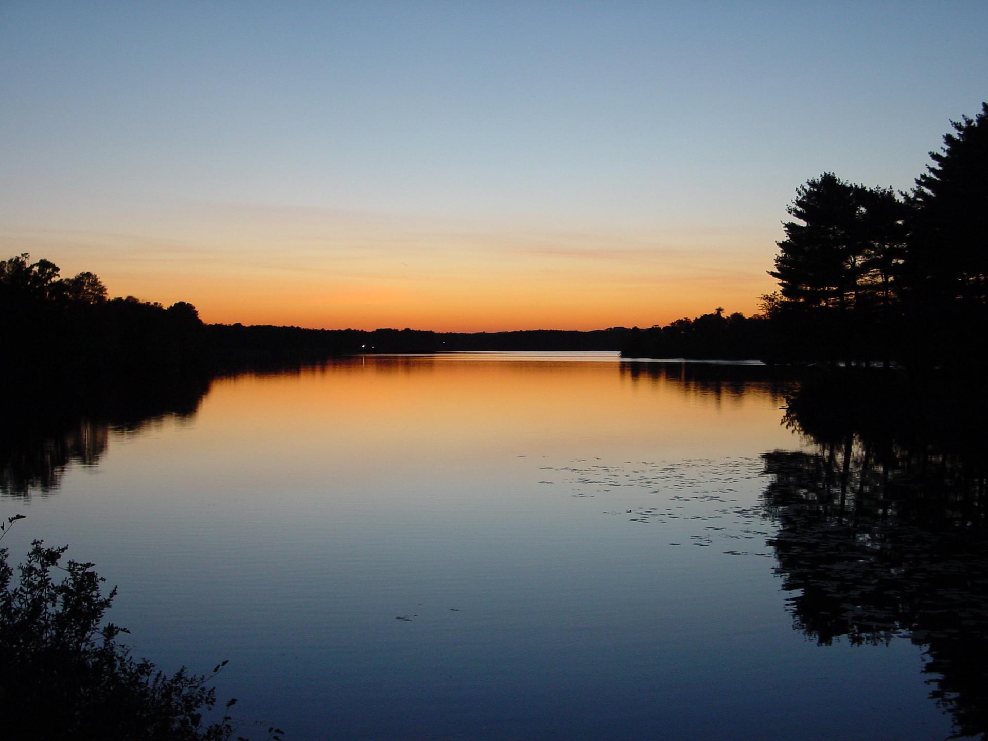 Silhouetted trees frame a tranquil lake reflecting a colorful sunset sky.