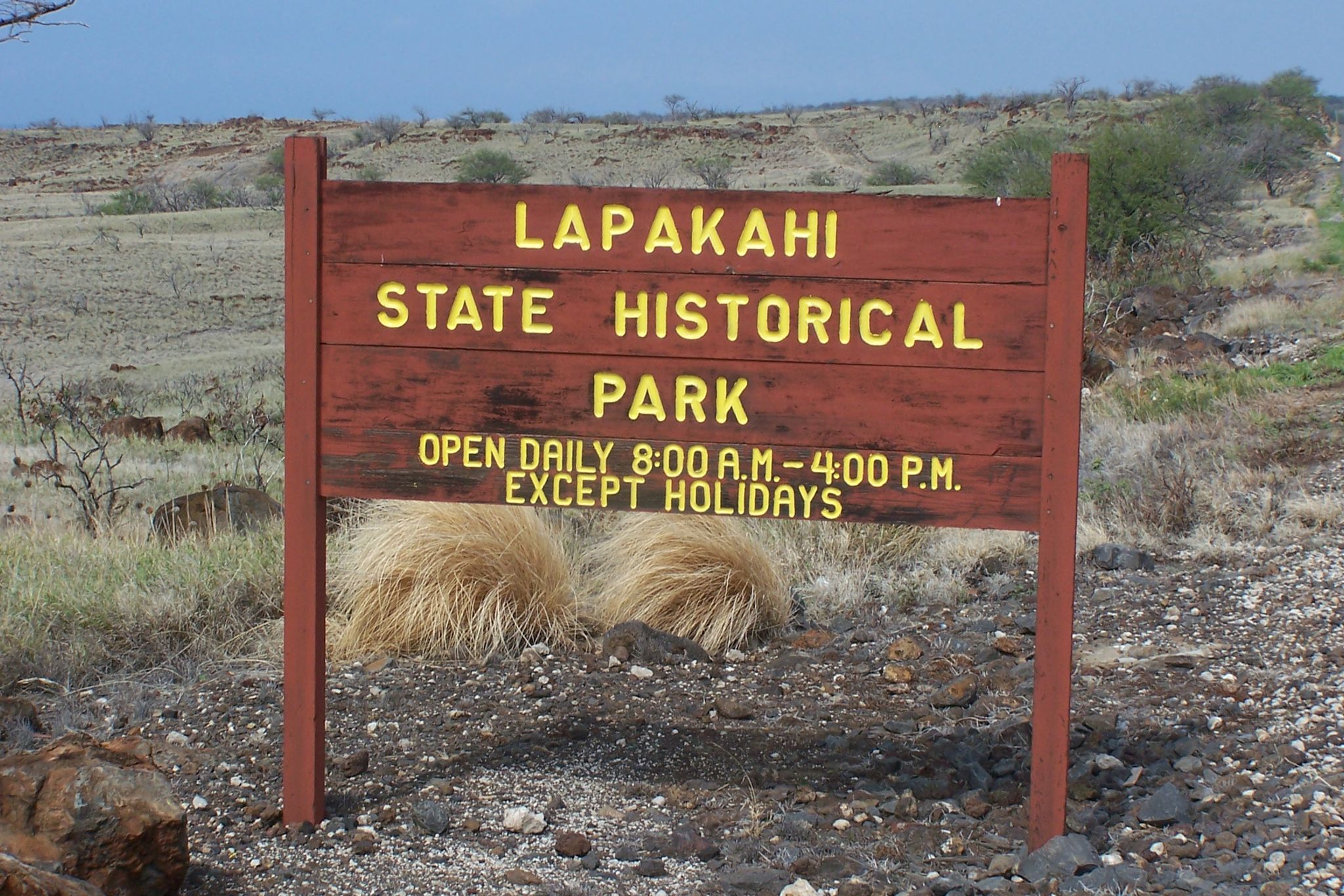 A weathered wooden sign stands in a dry, rocky landscape under a bright sky. - Akoni Pule Highway to Lapakahi State Historical Park