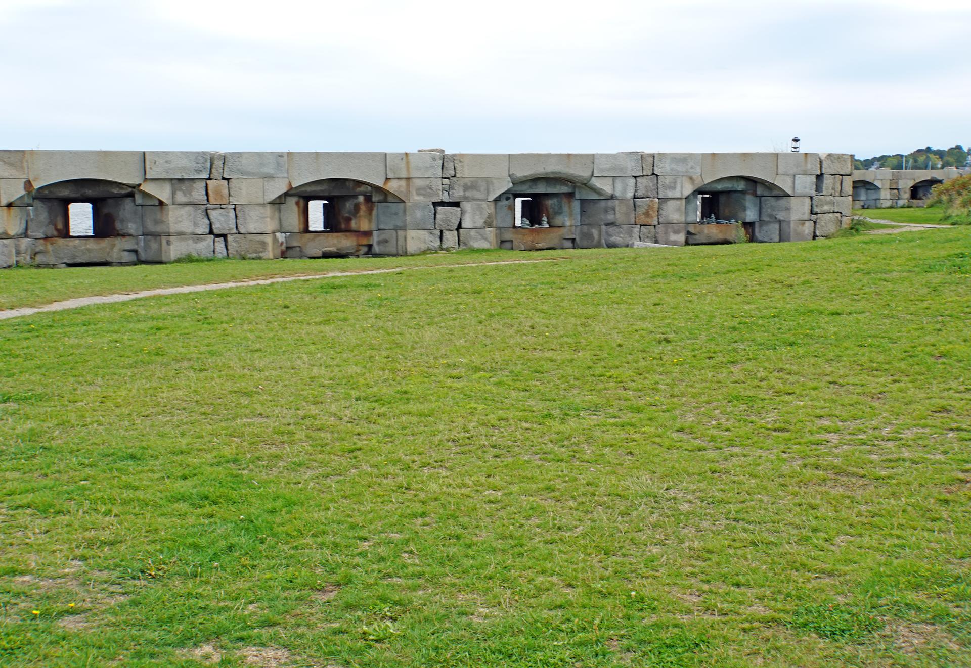 Long stone wall with arched openings next to a green field.