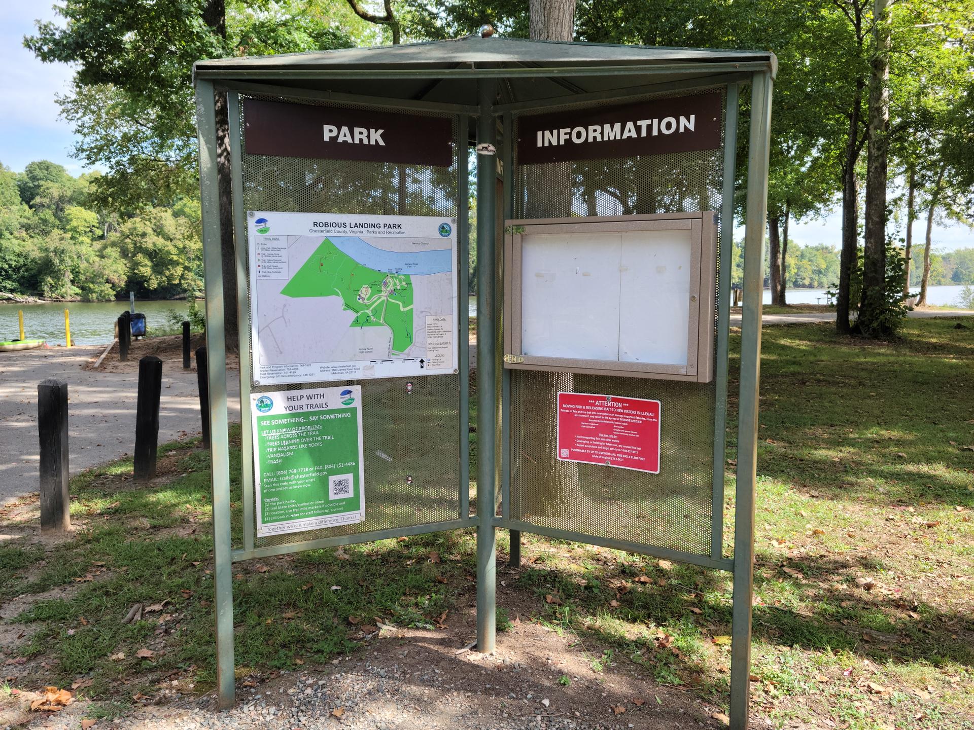 Park information kiosk with maps and notices, green trees, bright daylight.