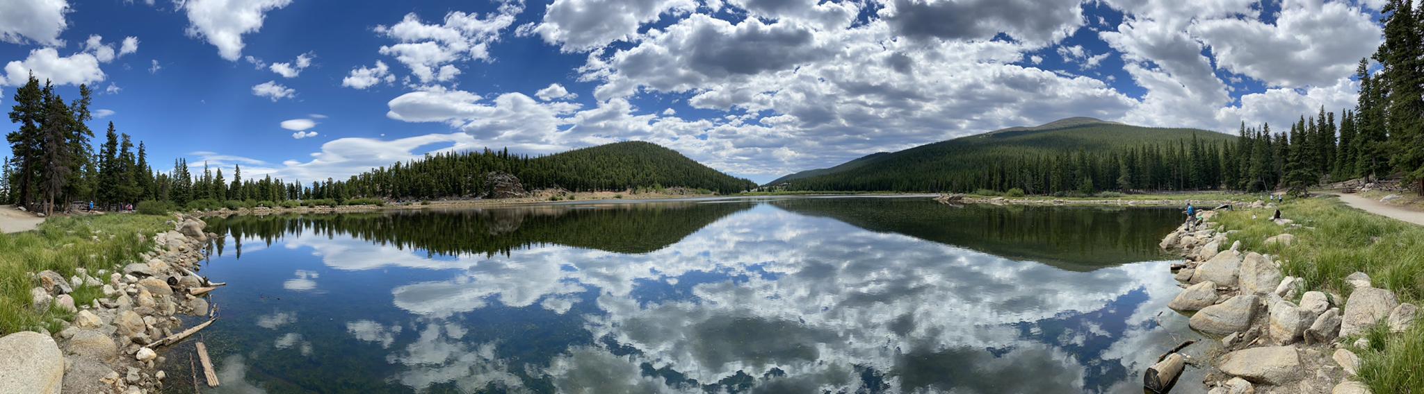Calm lake perfectly reflecting a very cloudy blue sky, surrounded by forested hills.
