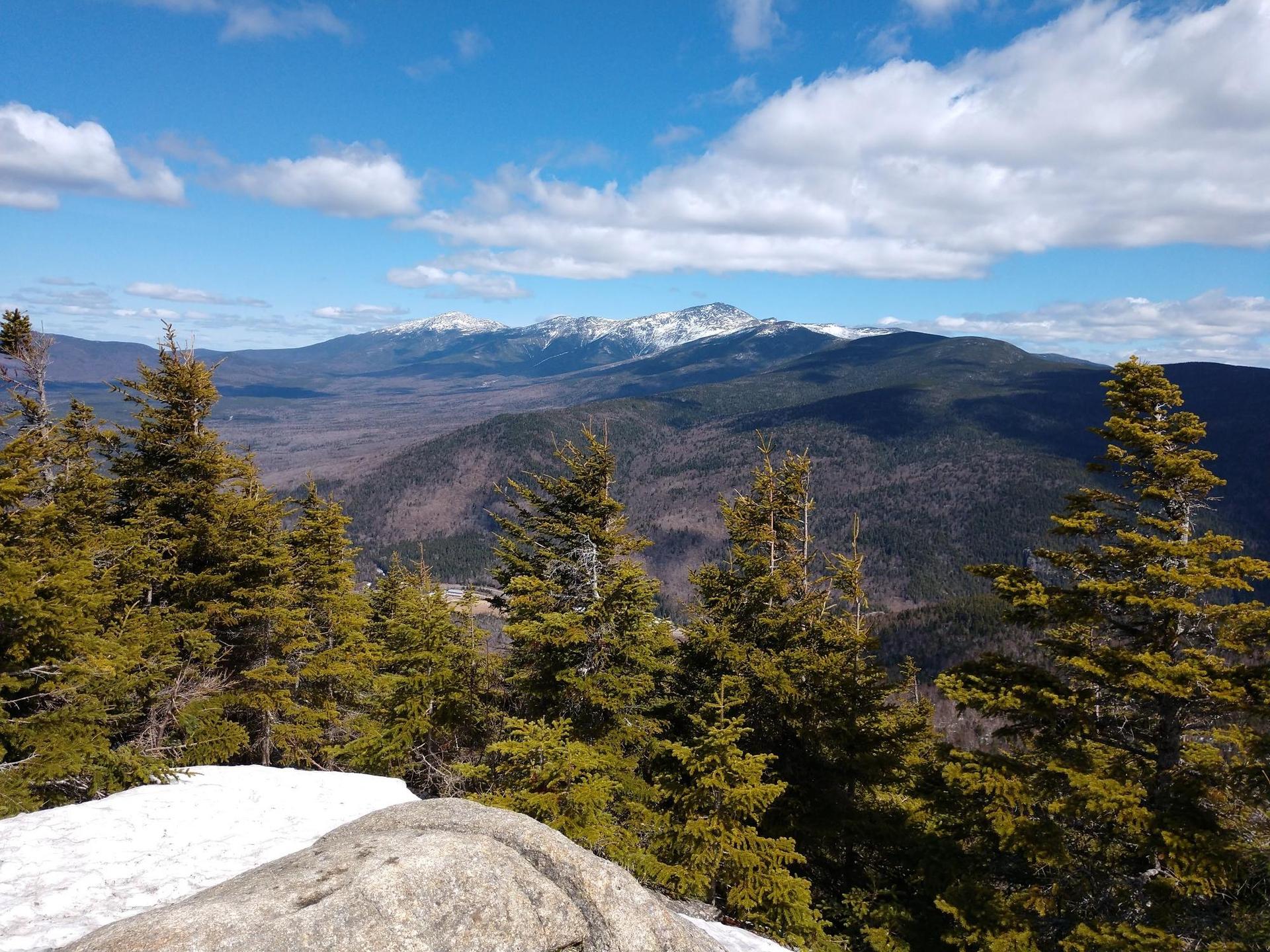 Snowy mountain peaks under a blue sky with clouds, seen through pine trees.