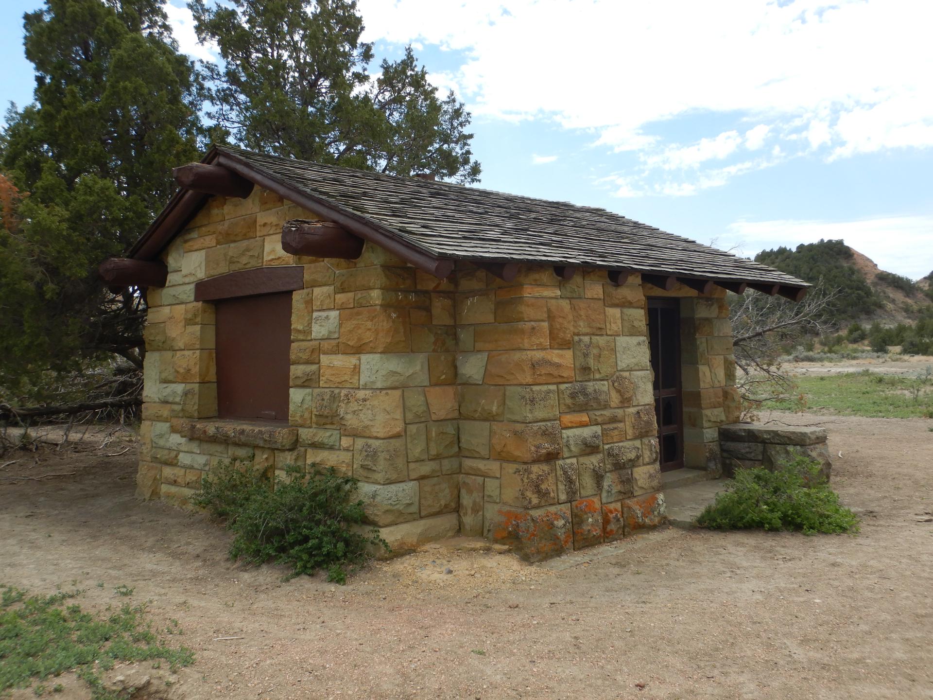 Stone building with a dark roof and shuttered window in a dry, sunny landscape.
