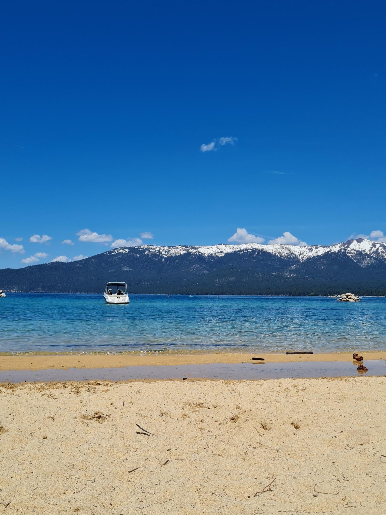 Sandy beach leading to a blue lake with a boat and snowy mountains beyond.