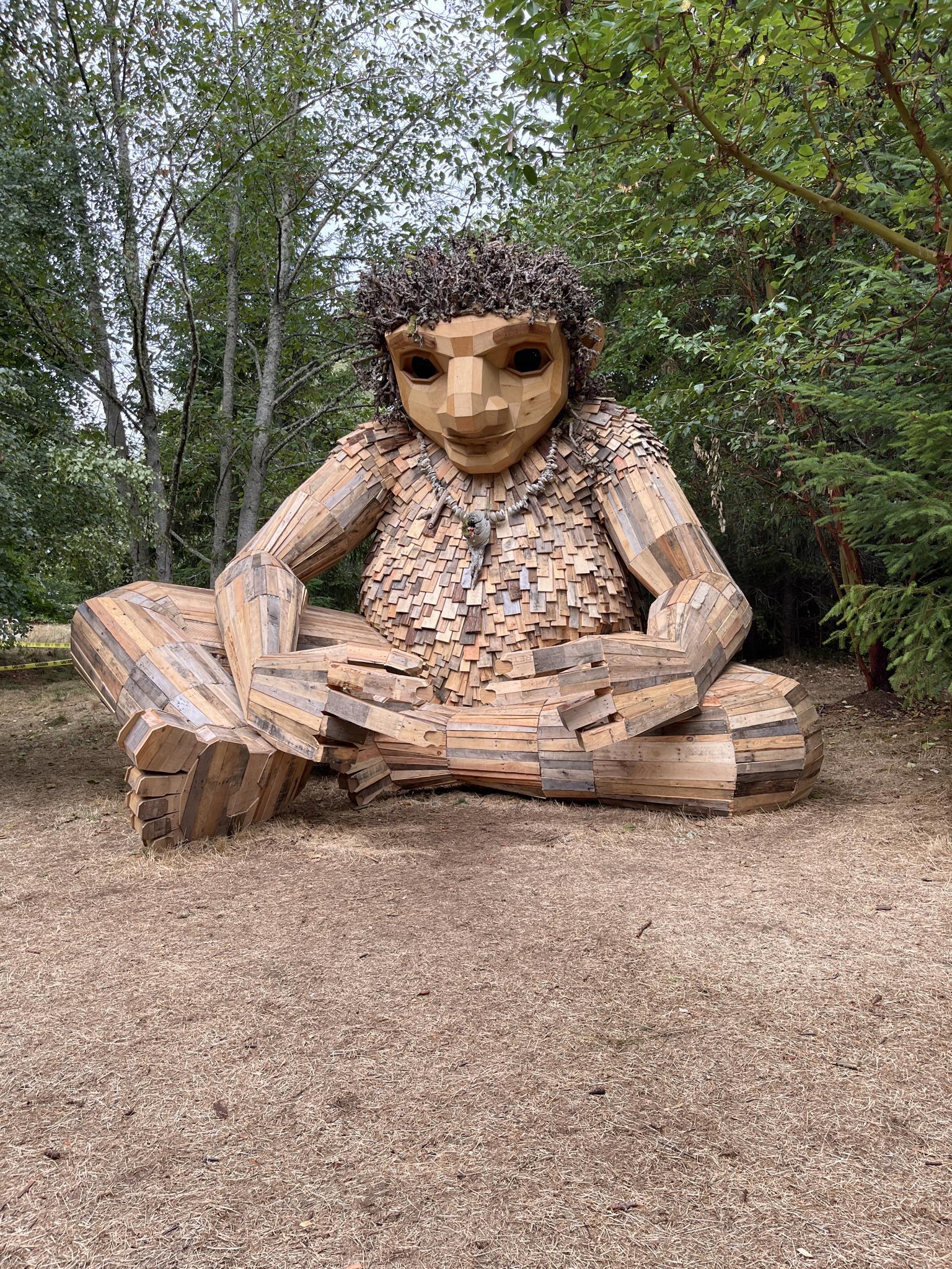 Large wooden sculpture of a seated figure in a forest clearing, bright light.