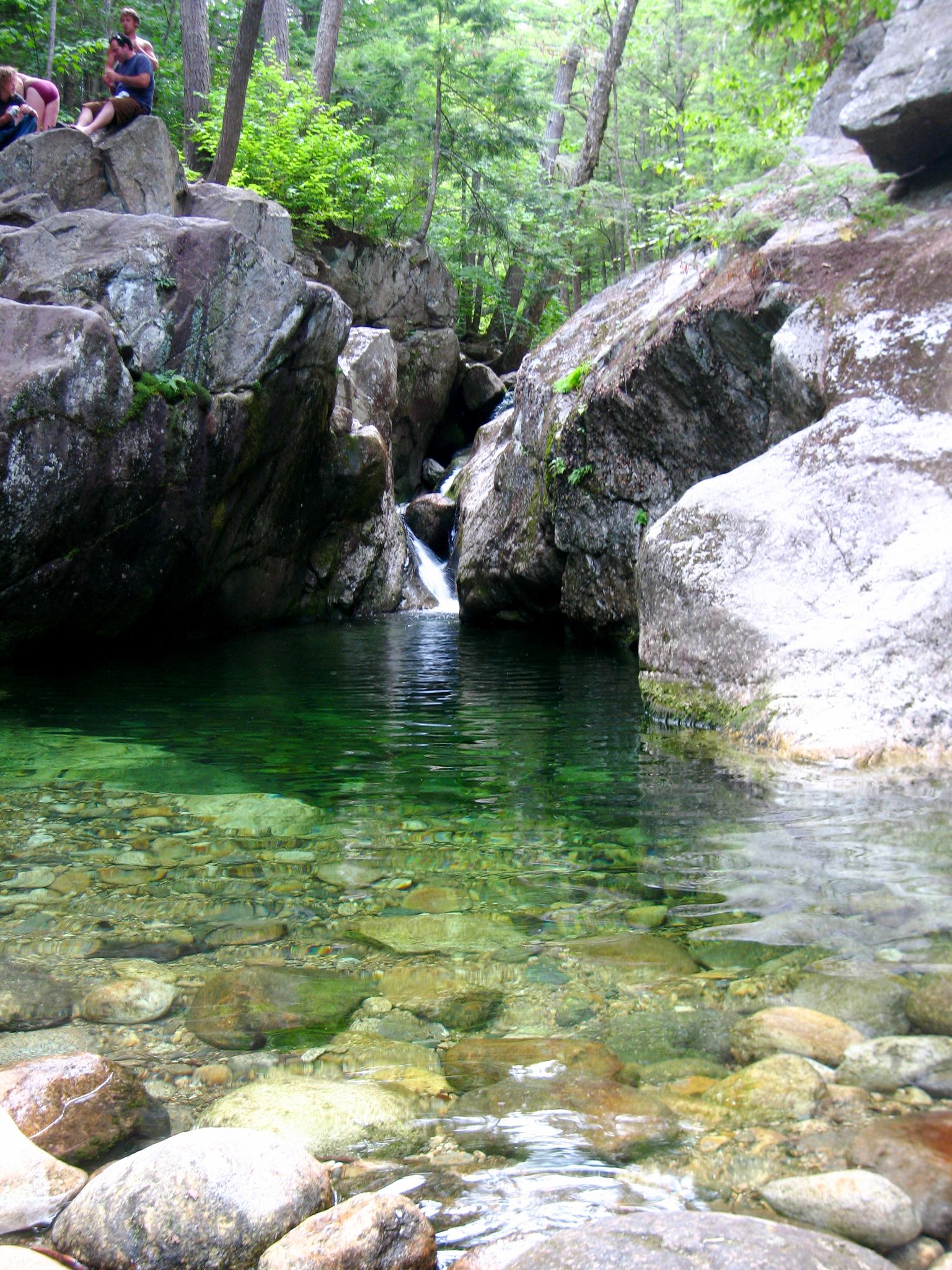People sit by a clear green pool with a waterfall and large rocks. - Emerald Pool Trail