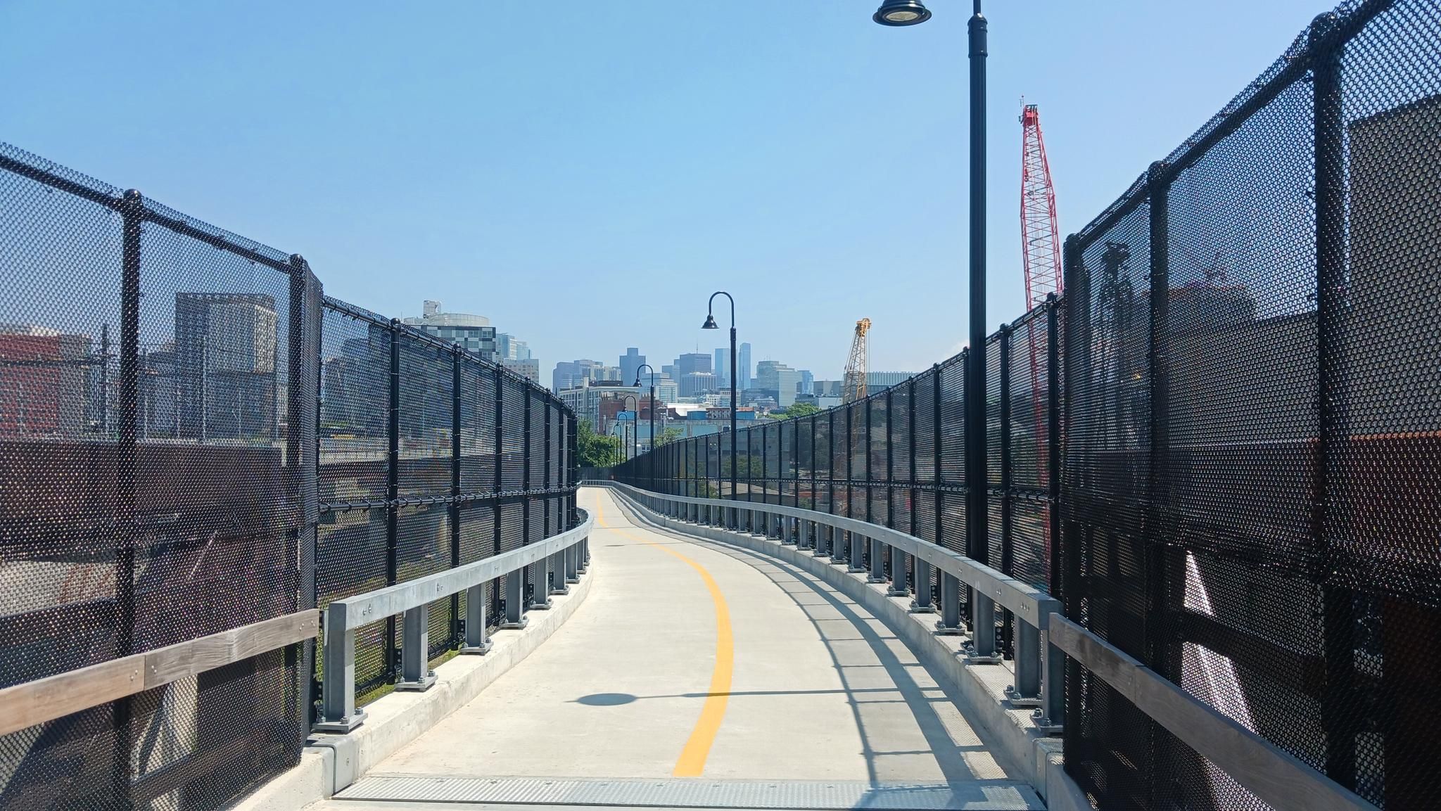 A fenced pedestrian pathway with a yellow line leading to a city. - Mass Central Rail Trail – Black Lives Matter graffiti loop from Somerville