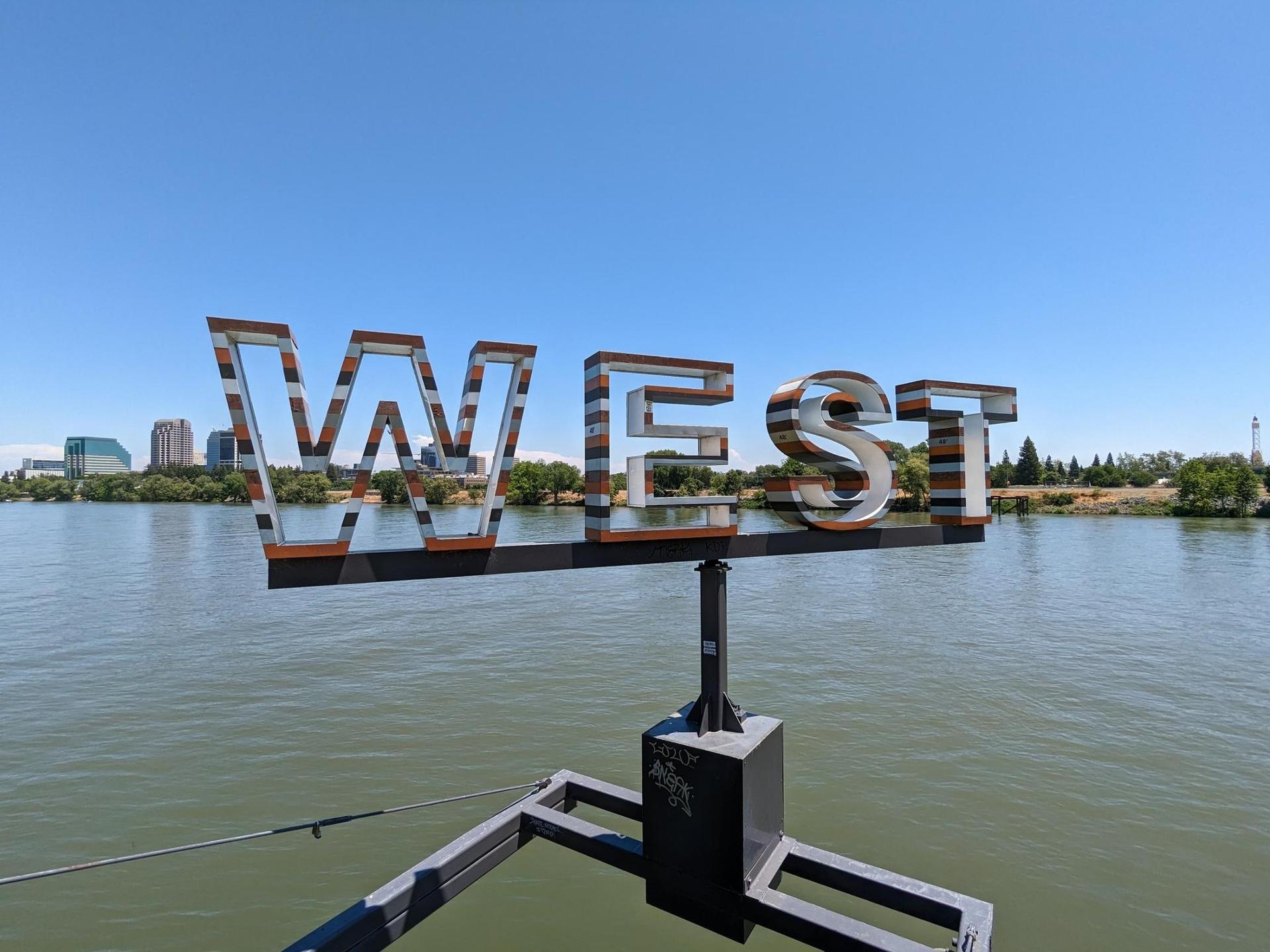 Large metal 'WEST' sign overlooking a wide river on a clear day.