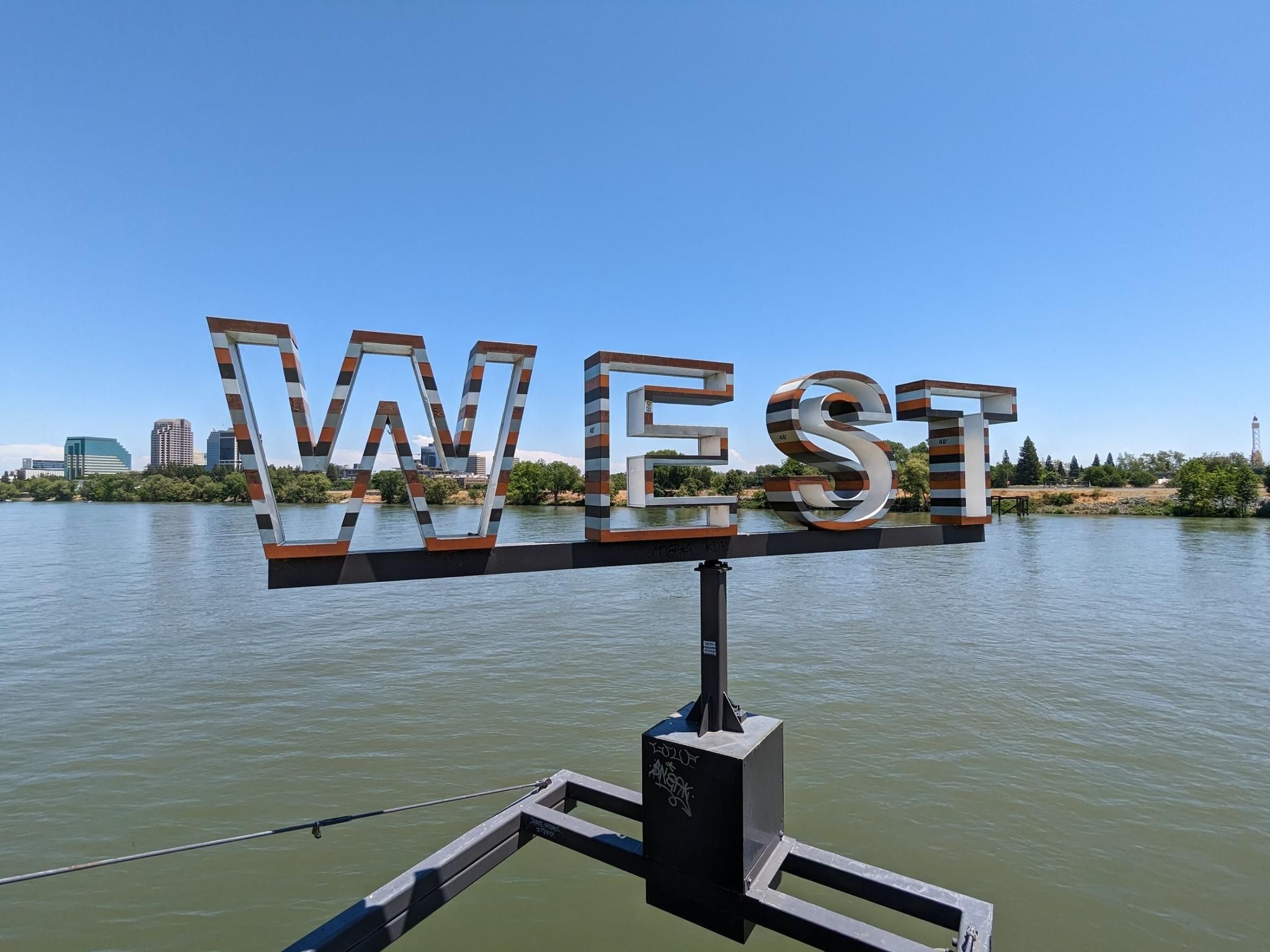 Large metal 'WEST' sign overlooking a wide river on a clear day. - River Walk Trail