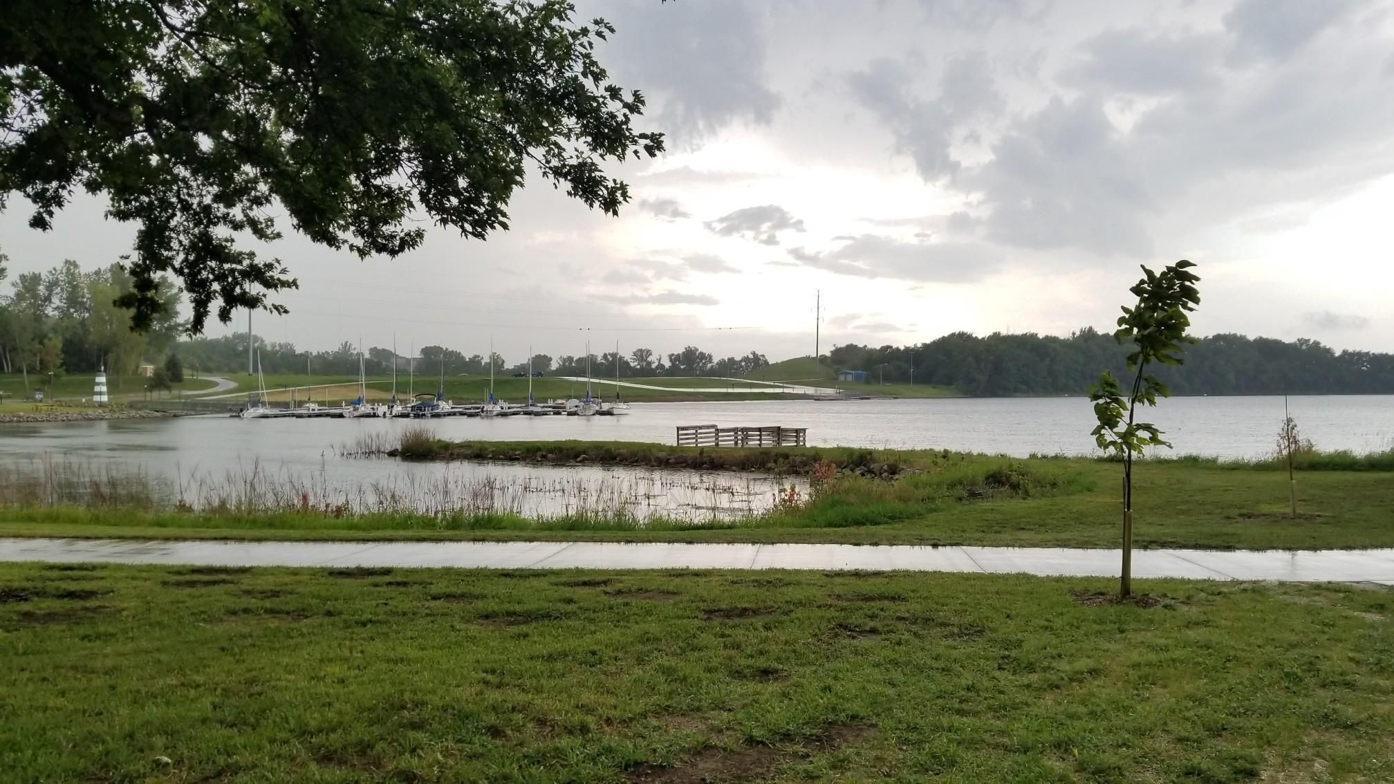 Overcast day over a lake with a marina, paved path, and green grass. - Cunningham Lake Pavilion – Glenn Cunningham Lake loop from Glenn Cunningham Lake Park