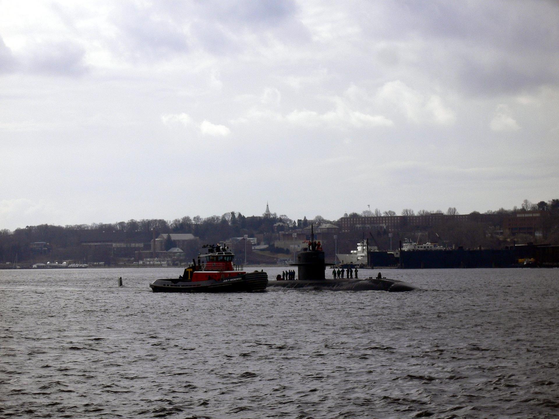 A tugboat guides a submarine on water under a cloudy sky.