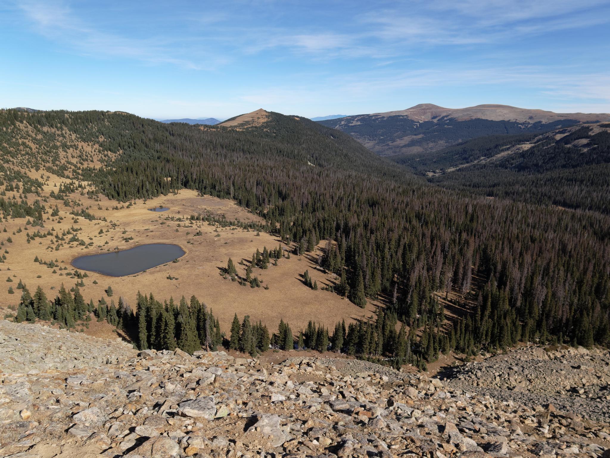 Rocky foreground overlooks a small lake, dense forests, and mountains under a clear sky.