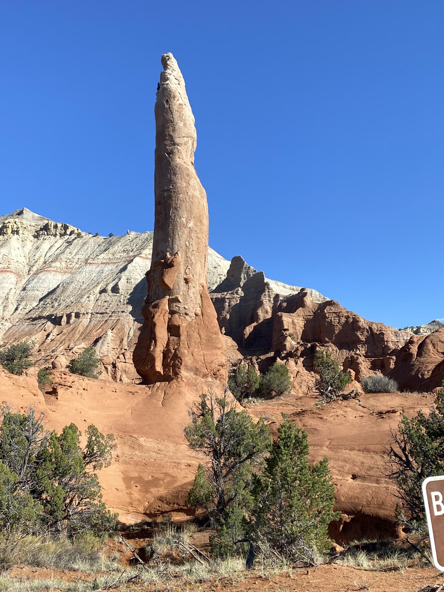 Tall, slender rock spire dominates a desert landscape under a clear blue sky.