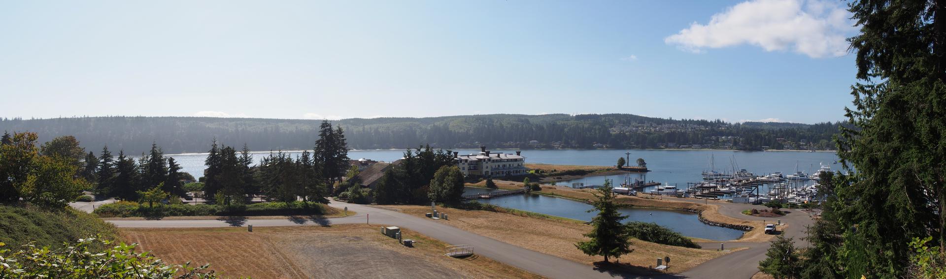 Wide shot of a sunny marina with boats, buildings, and distant forested land.