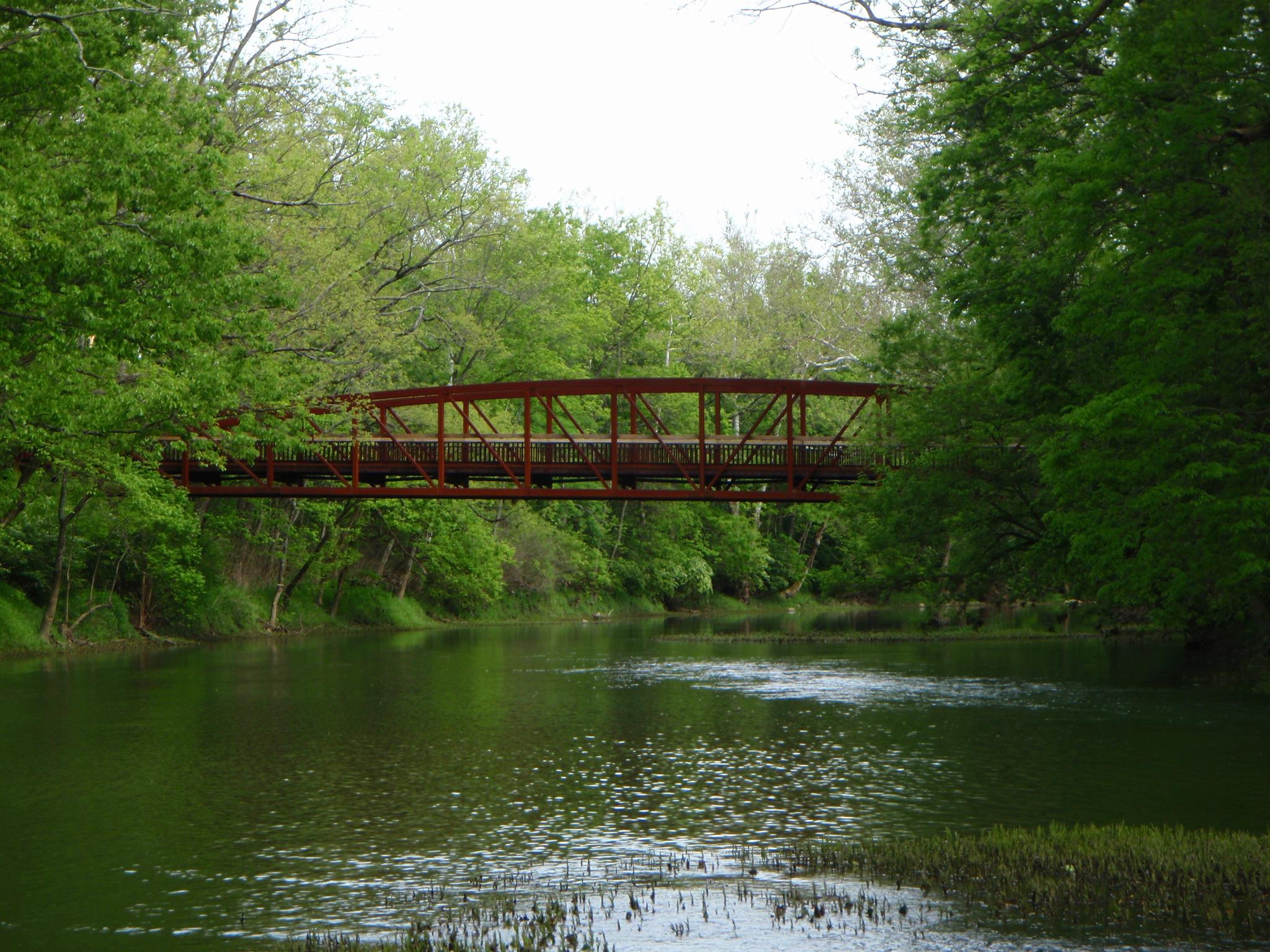 Red bridge spans a green river surrounded by lush trees under bright skies.