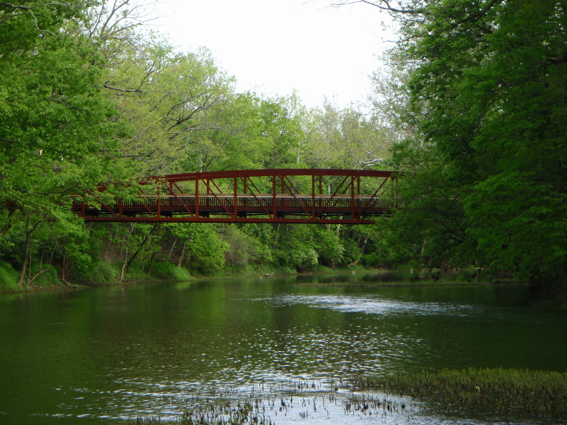 Red bridge spans a green river surrounded by lush trees under bright skies.
