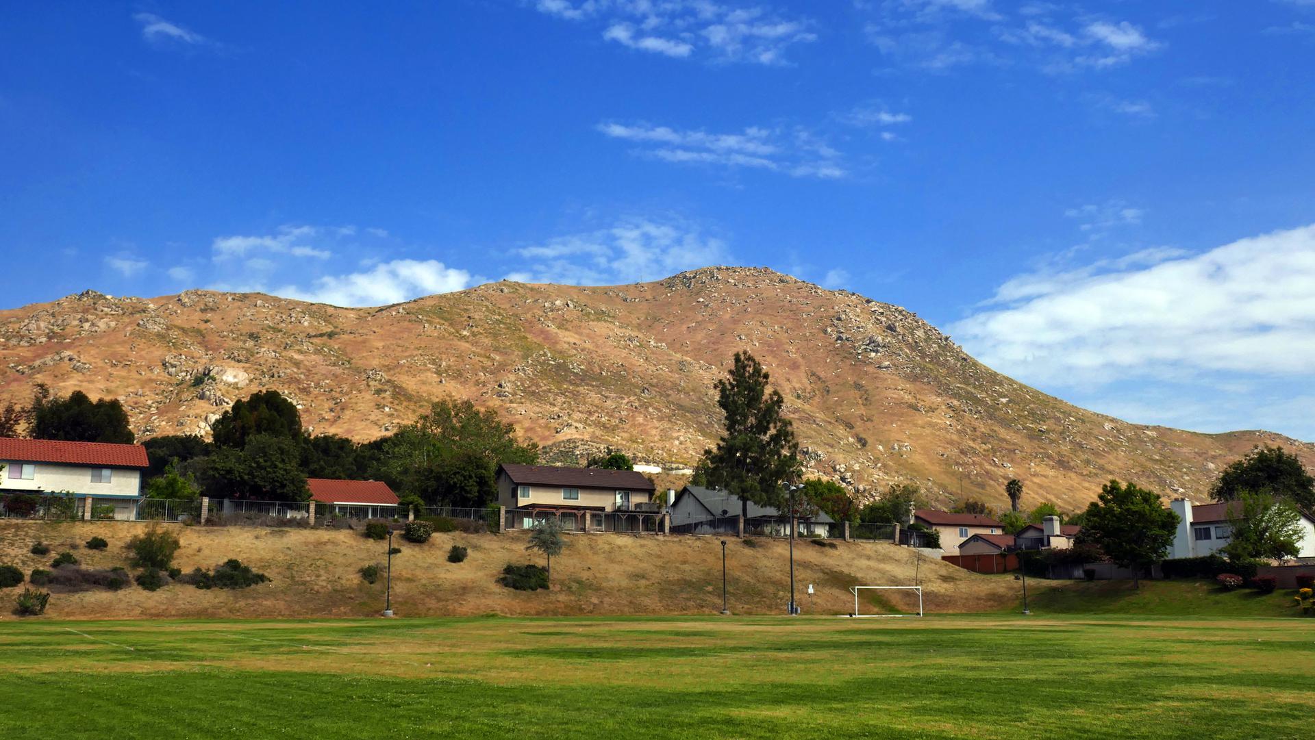 Green field, houses on a hillside, and a large dry mountain under blue sky.