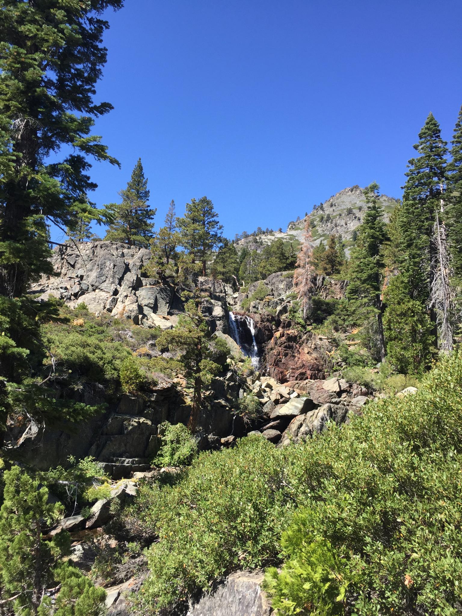 A waterfall cascades down a rocky, forested mountain under a clear blue sky.