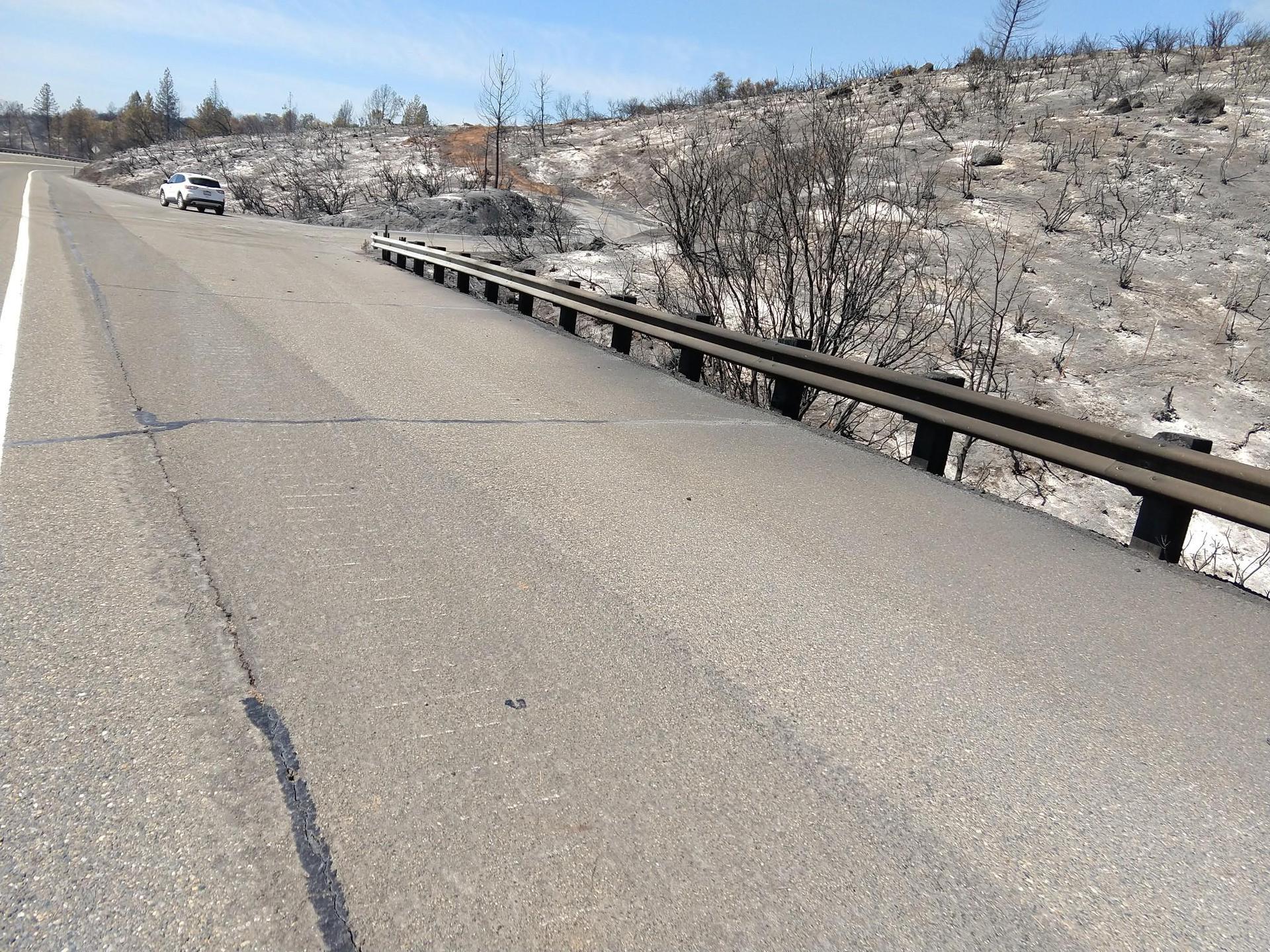 Asphalt road with guardrail next to a hillside of charred vegetation.