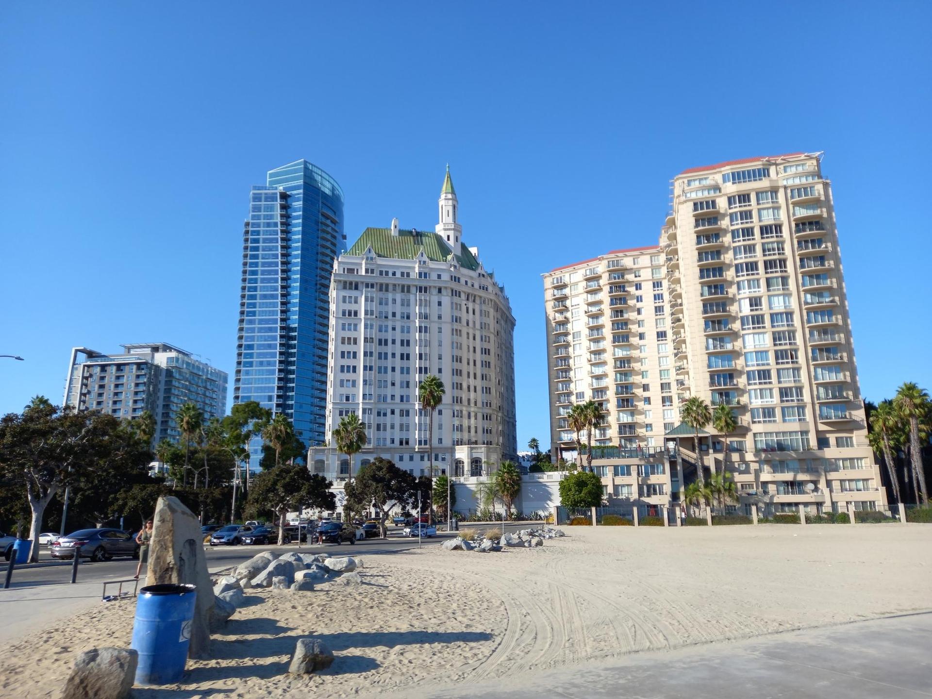 Tall city buildings rise behind a sandy area under a bright blue sky.