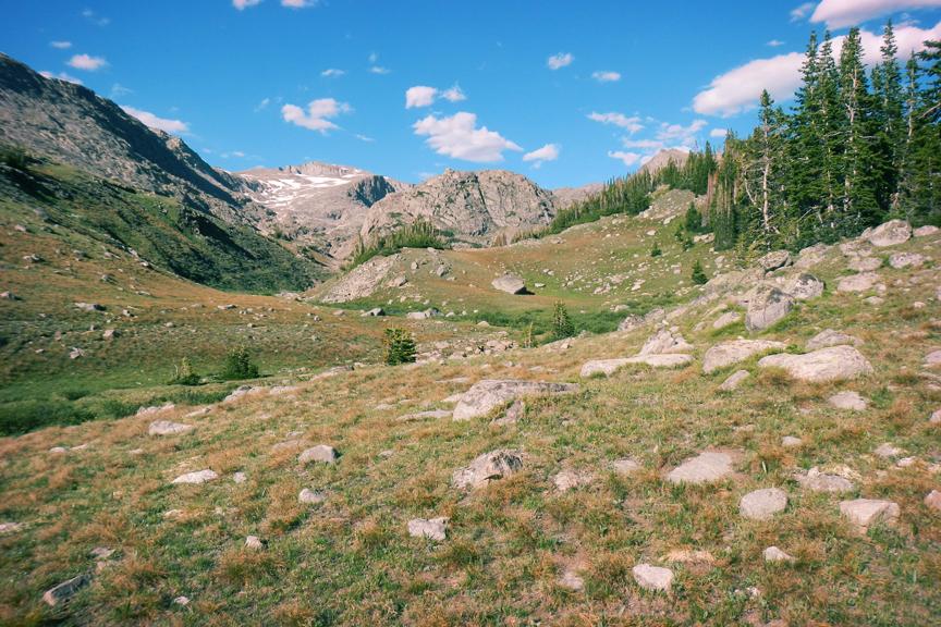 Expansive mountain valley with grassy slopes, rocky peaks, and scattered pines.