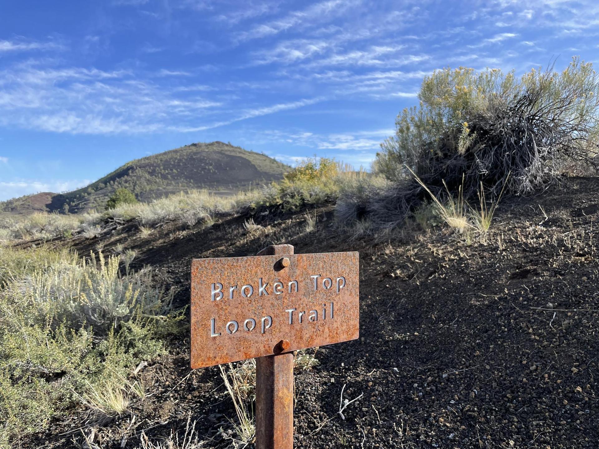 A rusty metal sign stands on a dry, rocky path under a blue sky.