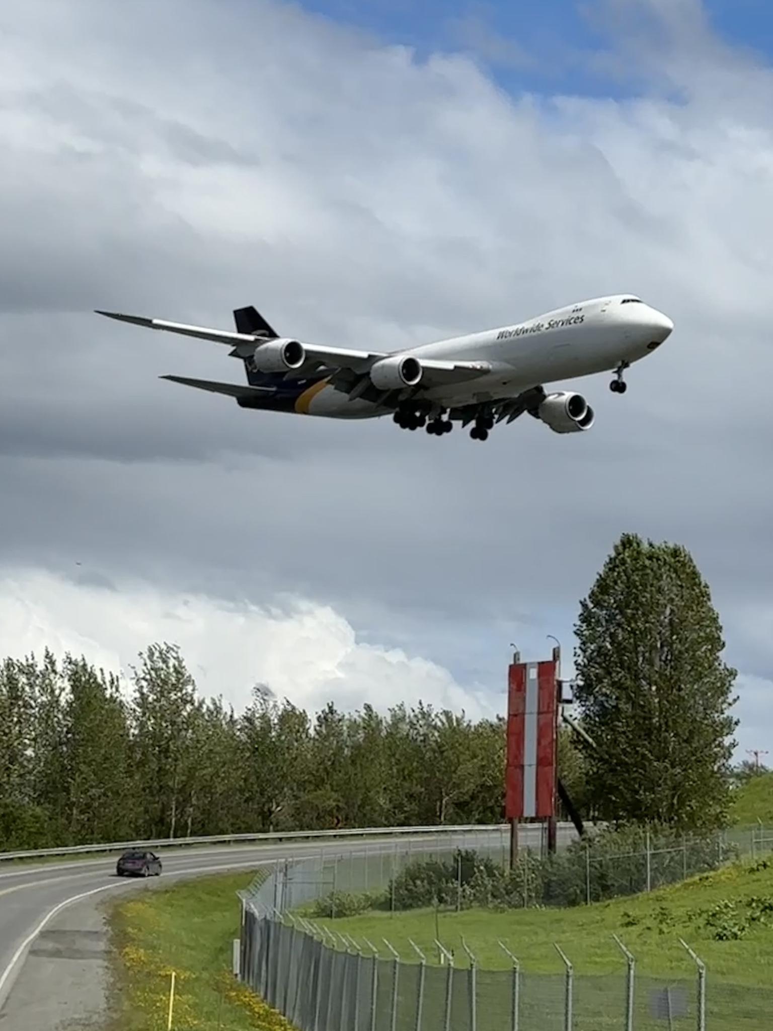 Large cargo airplane descending over a road and green trees under cloudy sky.