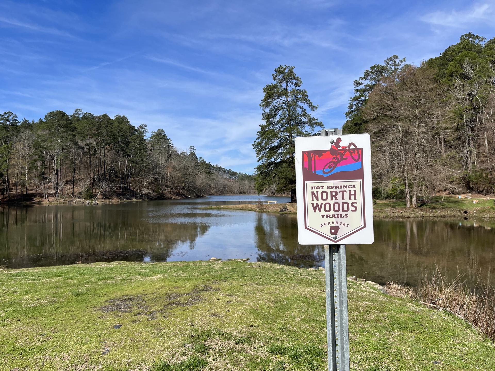 Trail sign by a calm lake surrounded by dense forest under a bright sky.